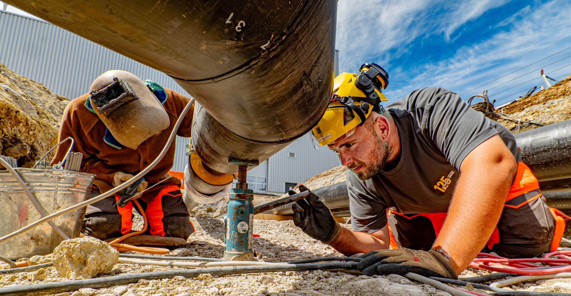 Ein Mann mit einem gelben Helm und Handschuhen führt Wartungsarbeiten an einer Rohrleitung durch, während ein Kollege im Hintergrund hilft.