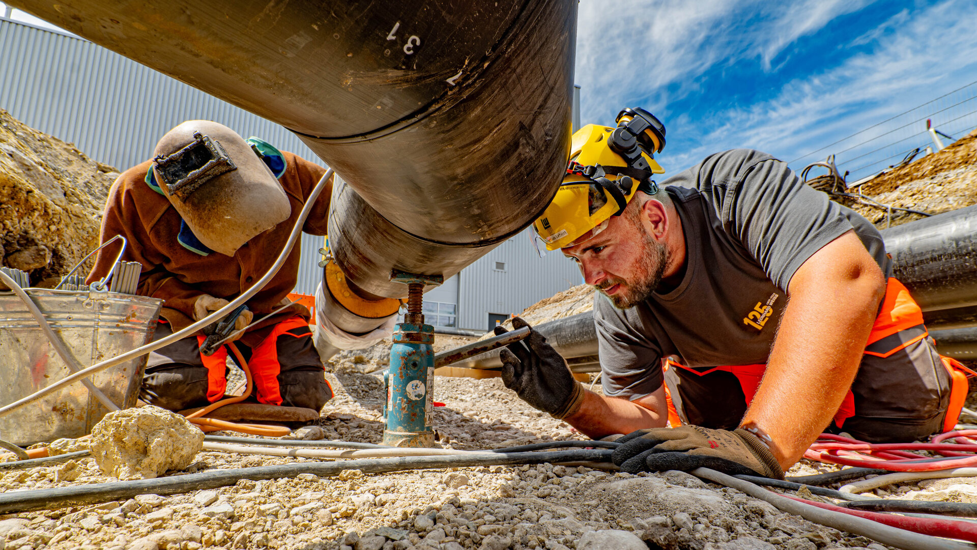 A man wearing a yellow helmet and gloves carries out maintenance work on a pipe while a colleague assists in the background.