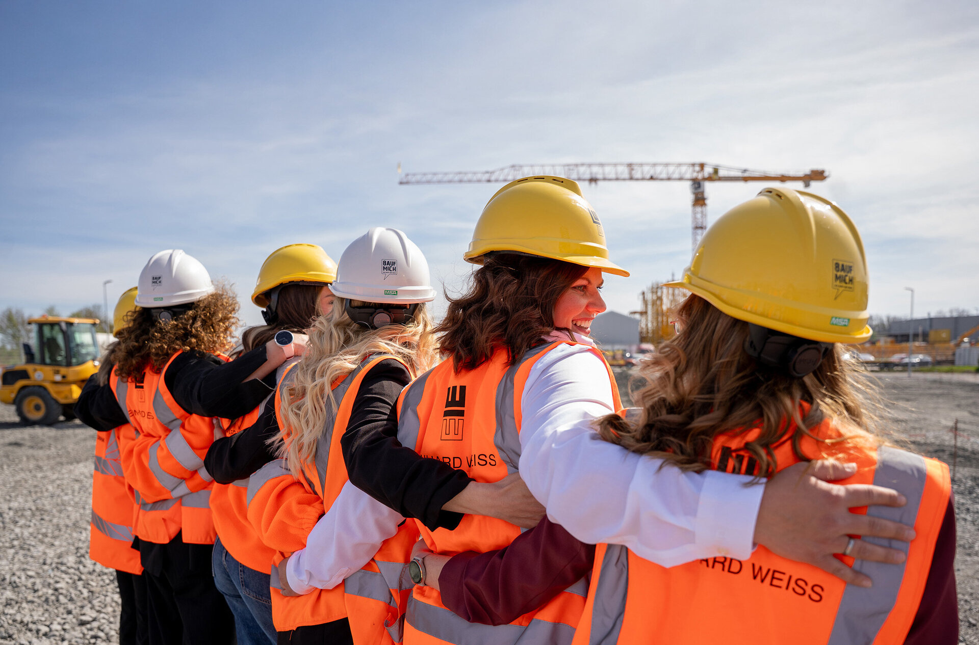 Eine Gruppe von Frauen in Sicherheitskleidung und Helmen umarmt sich, während sie auf eine Baustelle blicken.