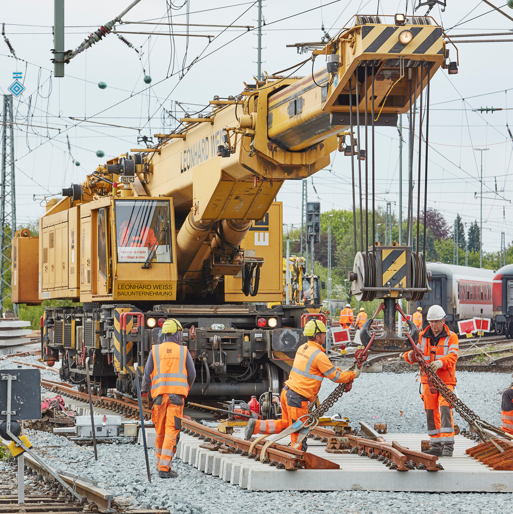 Ein Kran hebt Gleise, während mehrere Arbeiter in Sicherheitskleidung auf einer Bahnbaustelle aktiv sind.