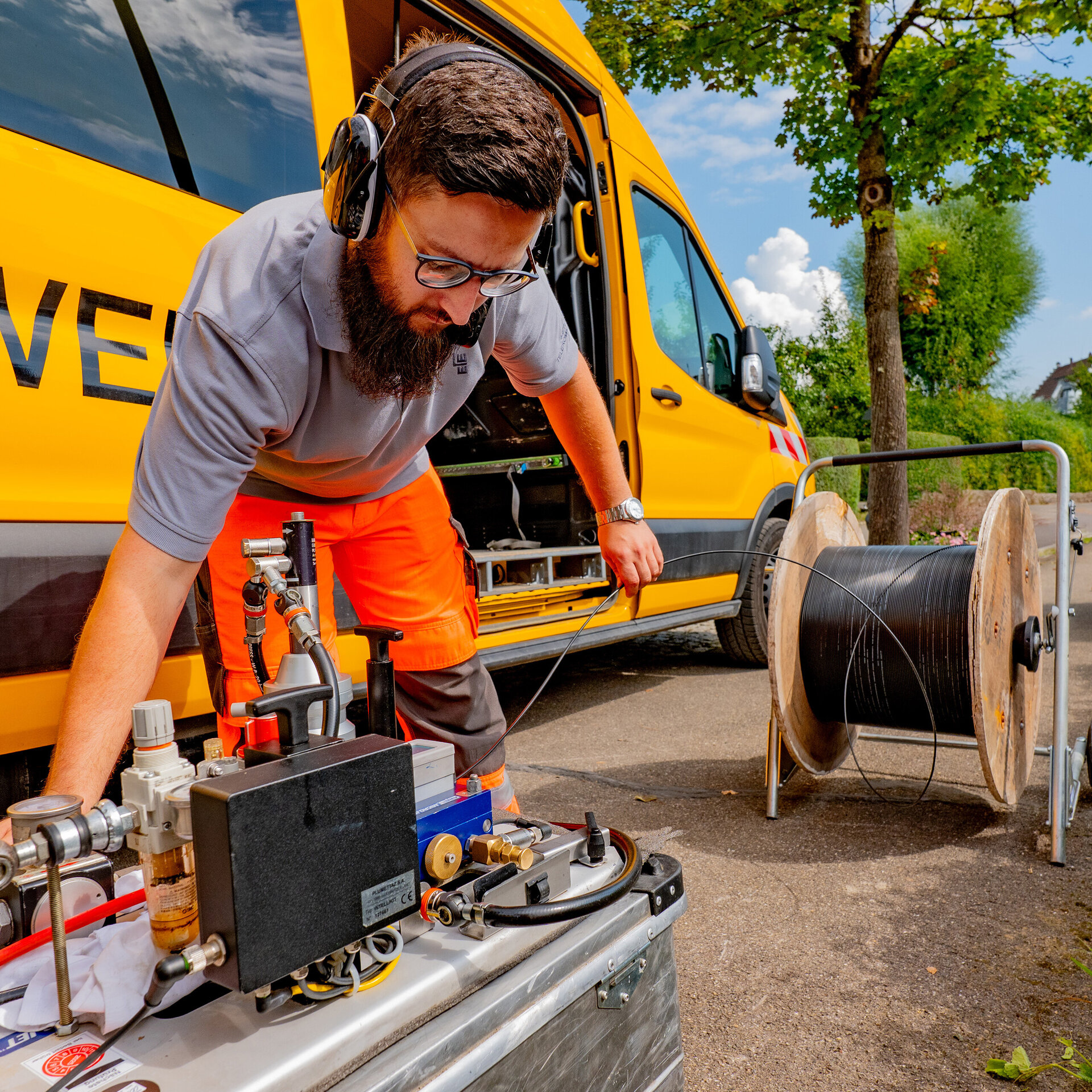 Ein Techniker in orangefarbener Arbeitskleidung arbeitet an einem Gerät neben einem gelben Fahrzeug auf einer Straße.