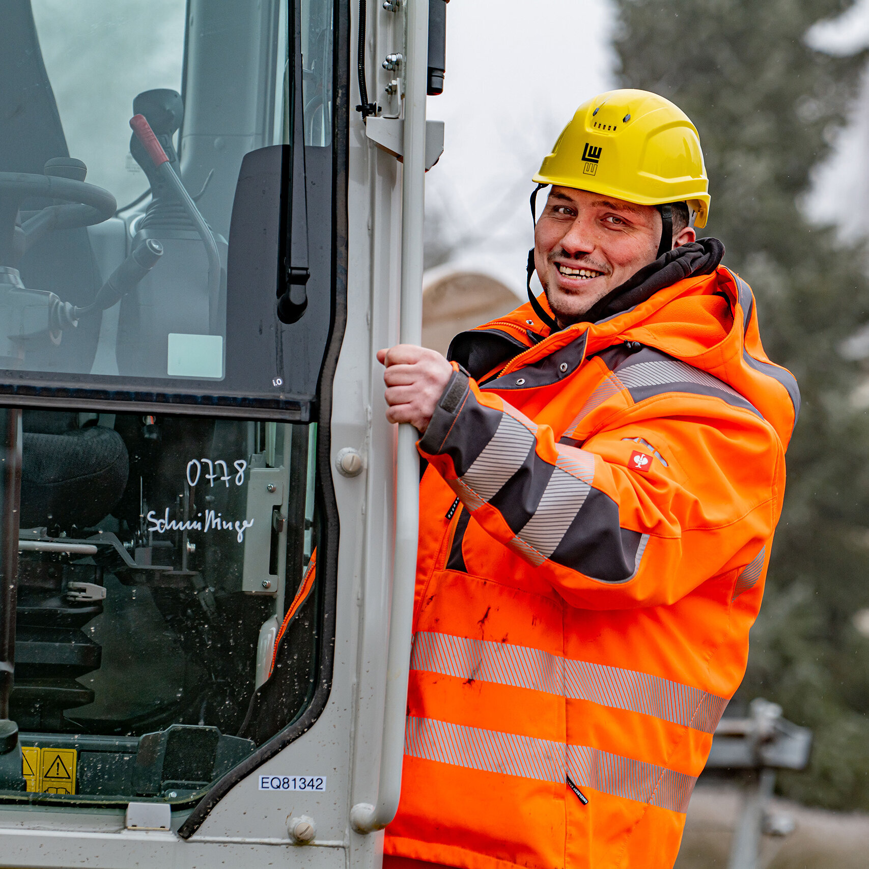 Ein lächelnder Arbeiter in leuchtend orangefarbener Sicherheitsjacke steht an einem Baumaschinenfahrzeug und begrüßt die Kamera.