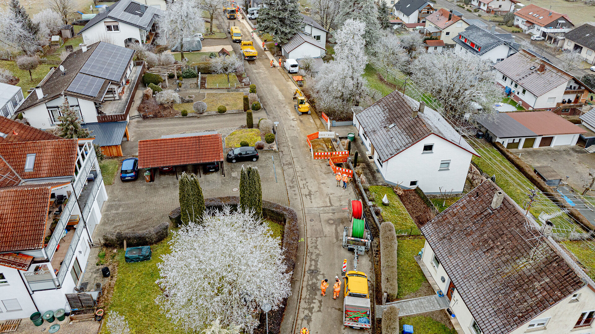 Blick auf eine ruhige Wohnstraße, wo Bauarbeiter mit orangefarbener Sicherheitskleidung an der Straße arbeiten, umgeben von frostigen Bäumen.