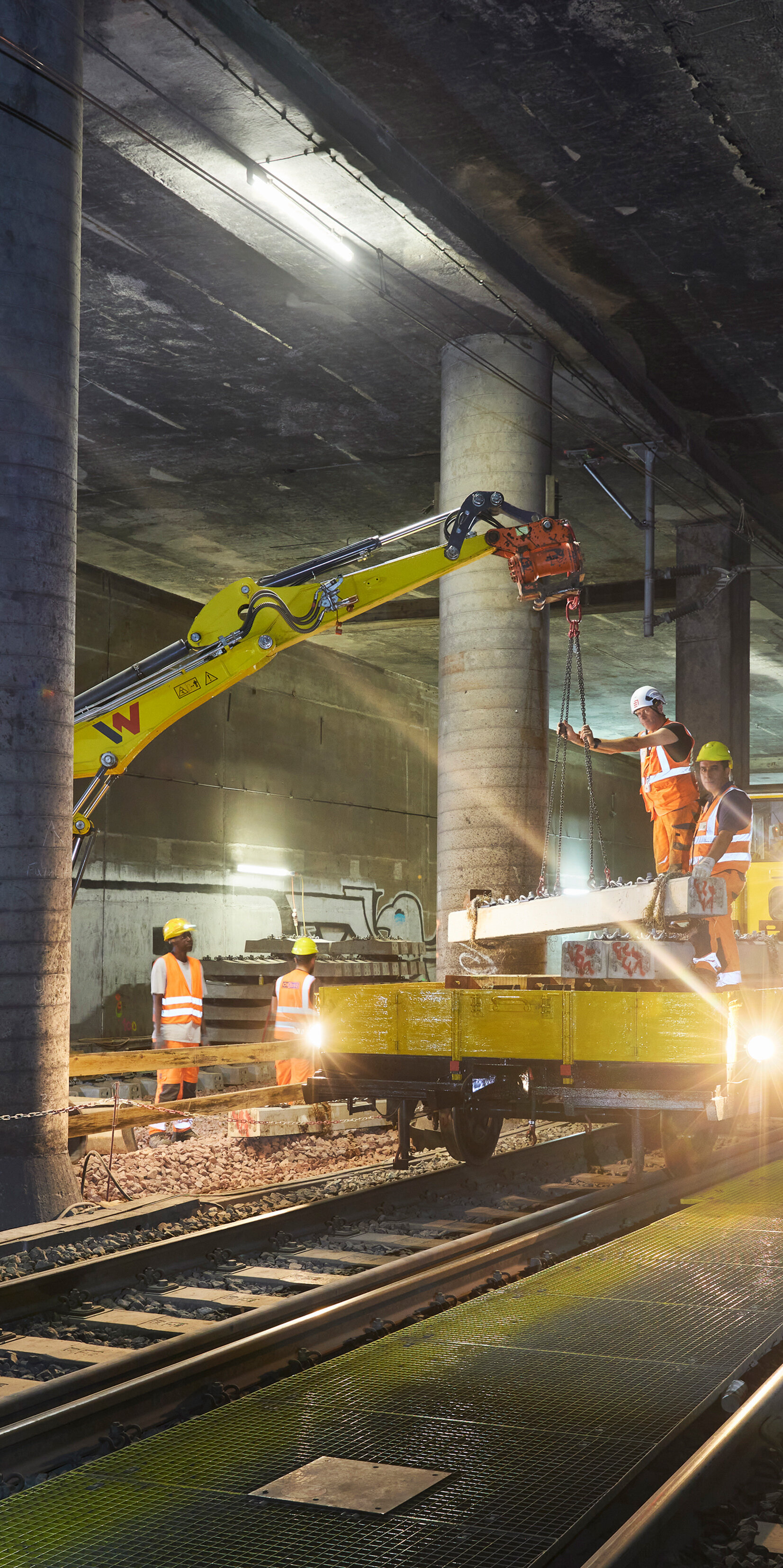 In einem unterirdischen Tunnel arbeiten Bauarbeiter an den Gleisen, umgeben von Betonpfeilern und schwachem Licht.