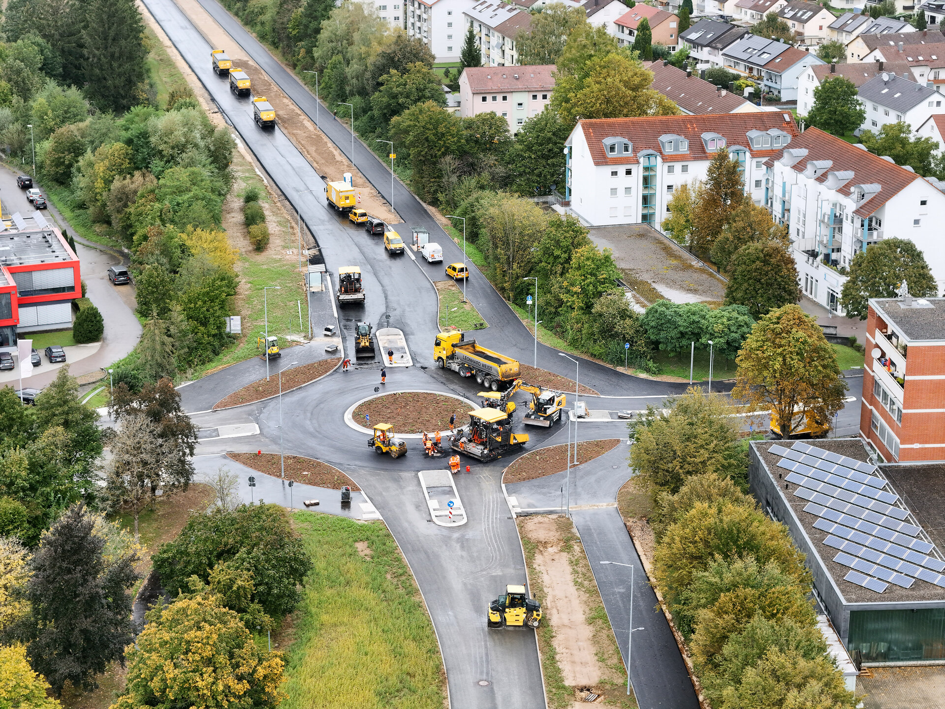 Eine Baustelle mit mehreren gelben Baumaschinen, die an einem neuen Kreisverkehr arbeiten, umgeben von Bäumen und Wohngebäuden.