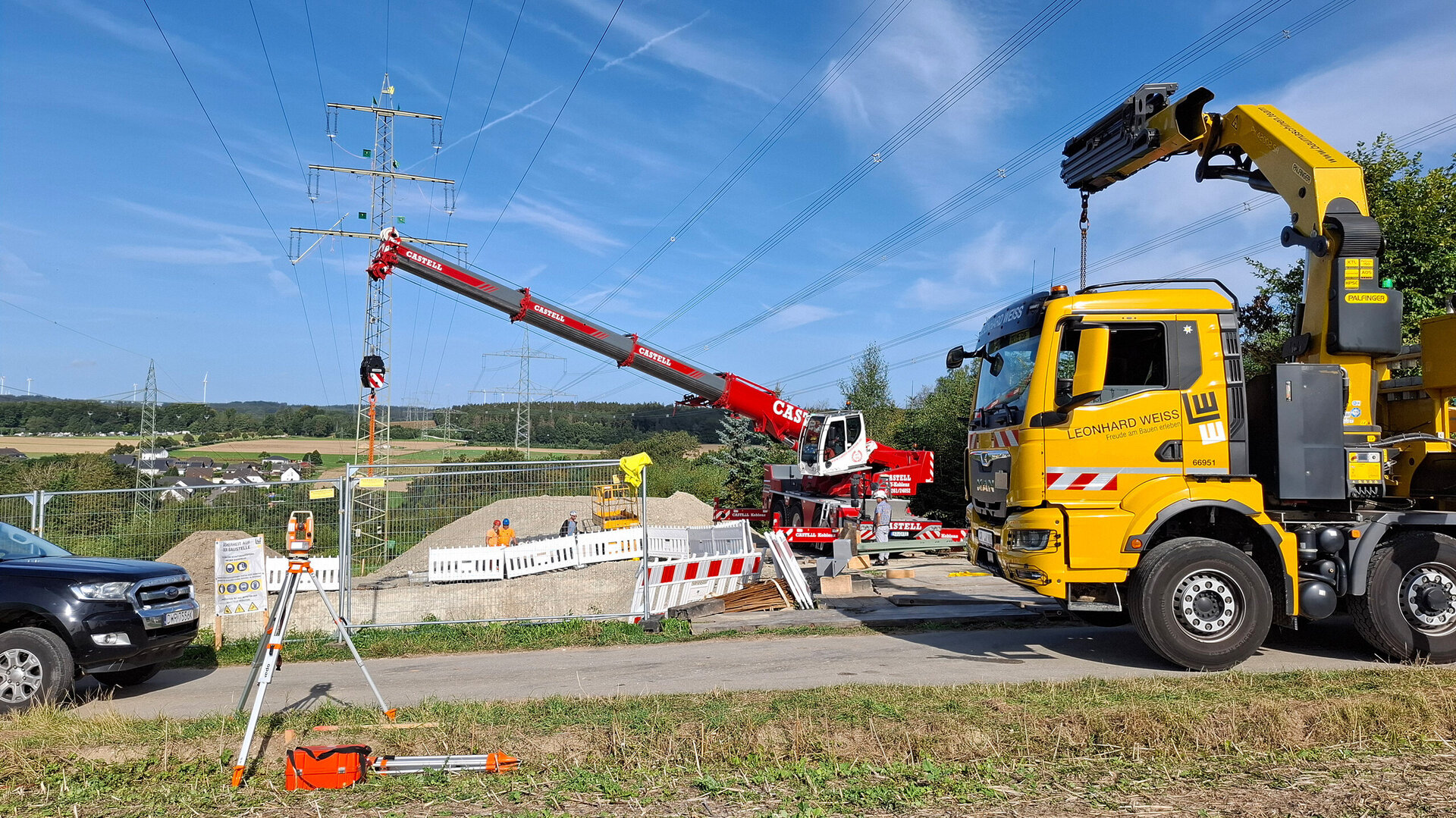 Baustellenaktivitäten sind im Gange, mit einem roten Kran, der an einem Strommast arbeitet, umgeben von Baugeräten und Arbeitern.