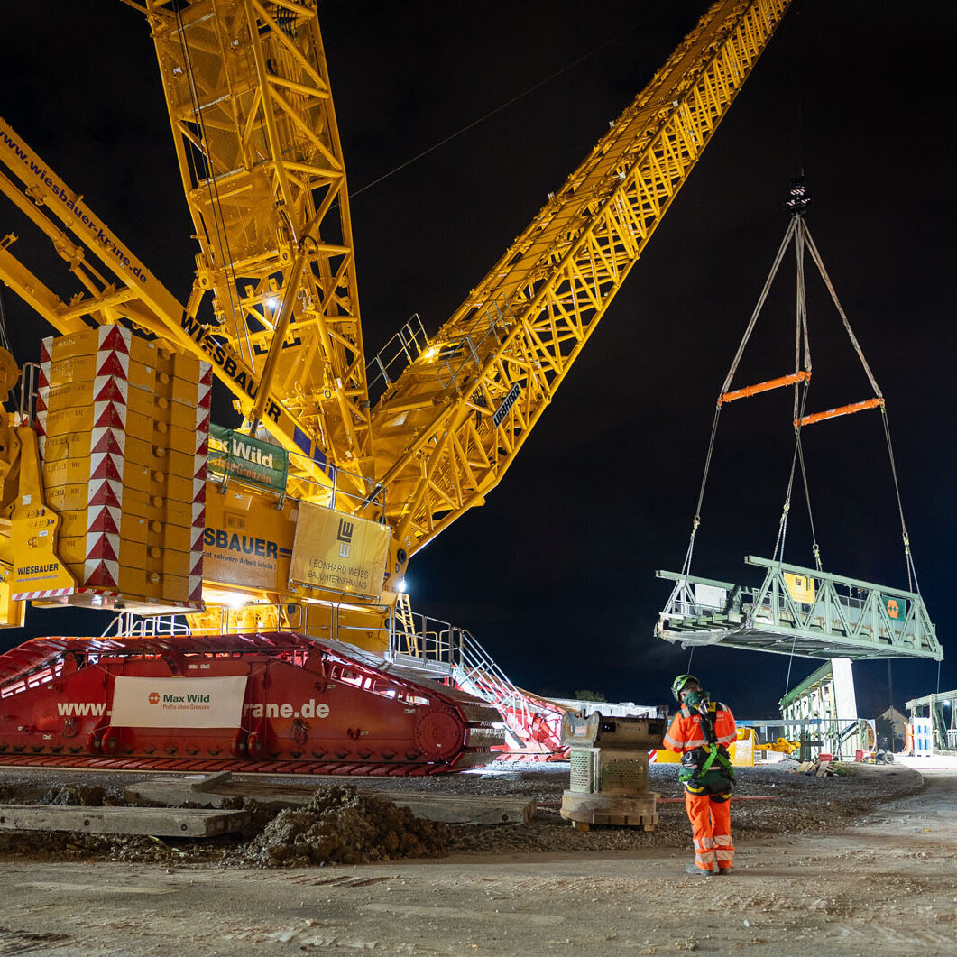 Ein großer gelber Kran hebt nachts eine massive Struktur, während Arbeiter in orangefarbener Sicherheitskleidung zuschauen.