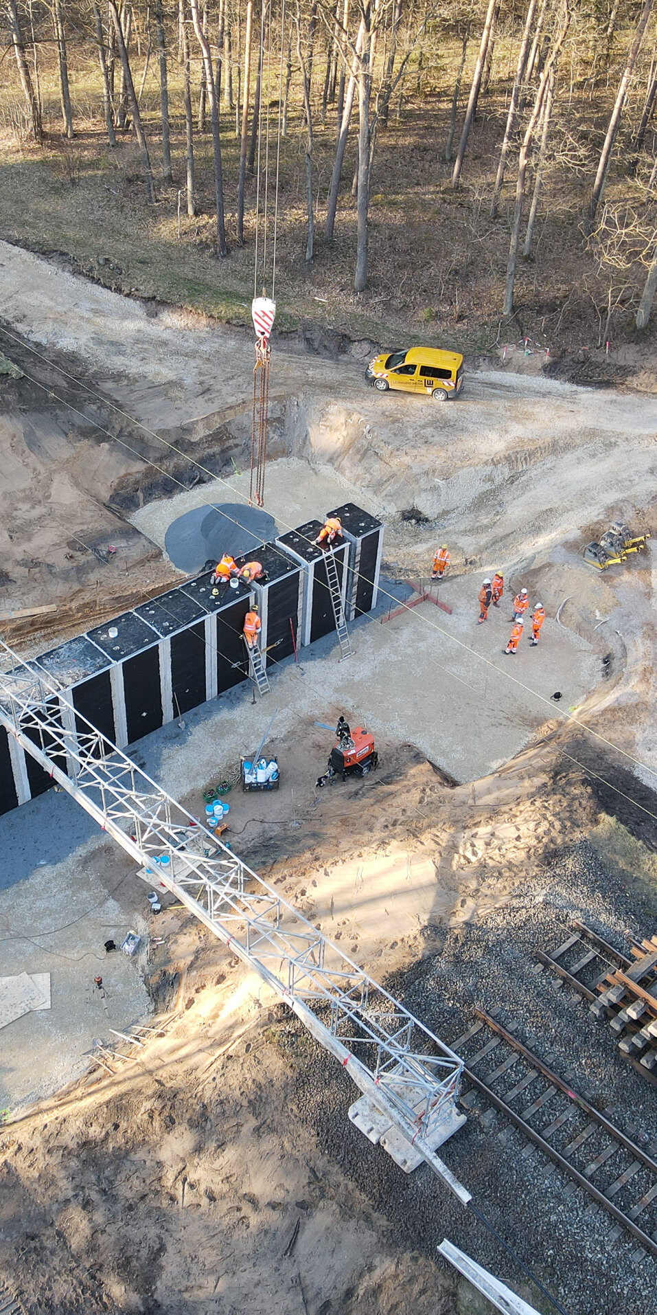 Die Szene zeigt eine Baustelle mit einem Kran, Bauarbeitern und einem unbefestigten Weg, umgeben von Bäumen und Gleisen.
