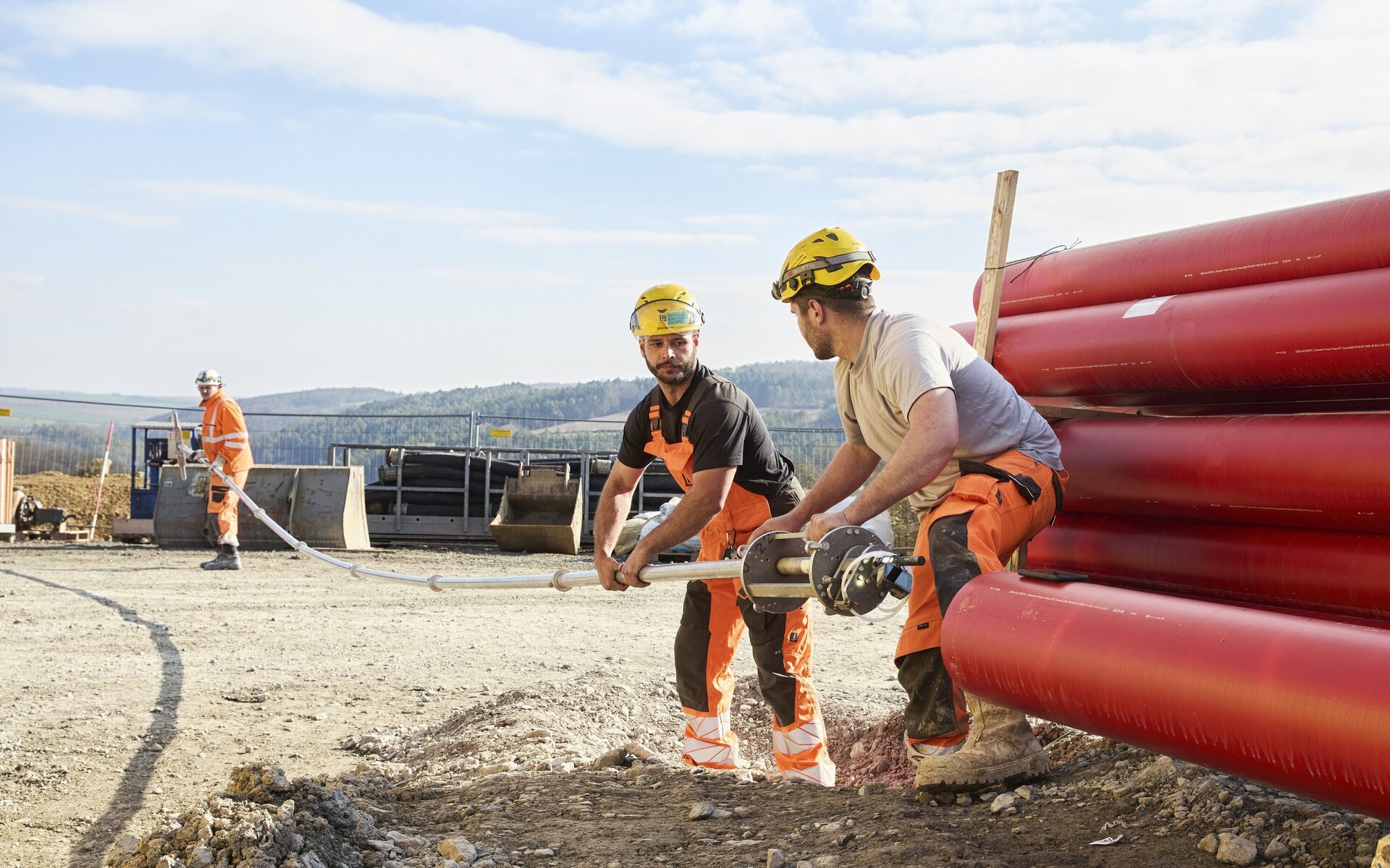 Die Szene zeigt zwei Bauarbeiter, die ein Rohr halten, während ein dritter Arbeiter im Hintergrund mit einem Werkzeug beschäftigt ist.