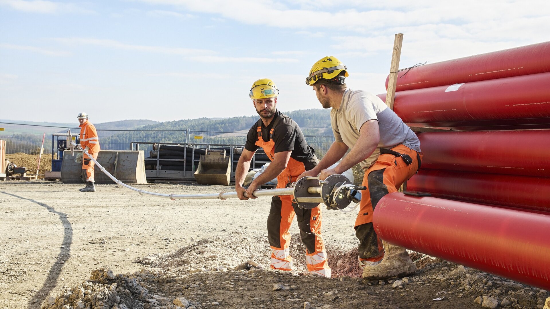 Die Szene zeigt zwei Bauarbeiter, die ein Rohr halten, während ein dritter Arbeiter im Hintergrund mit einem Werkzeug beschäftigt ist.