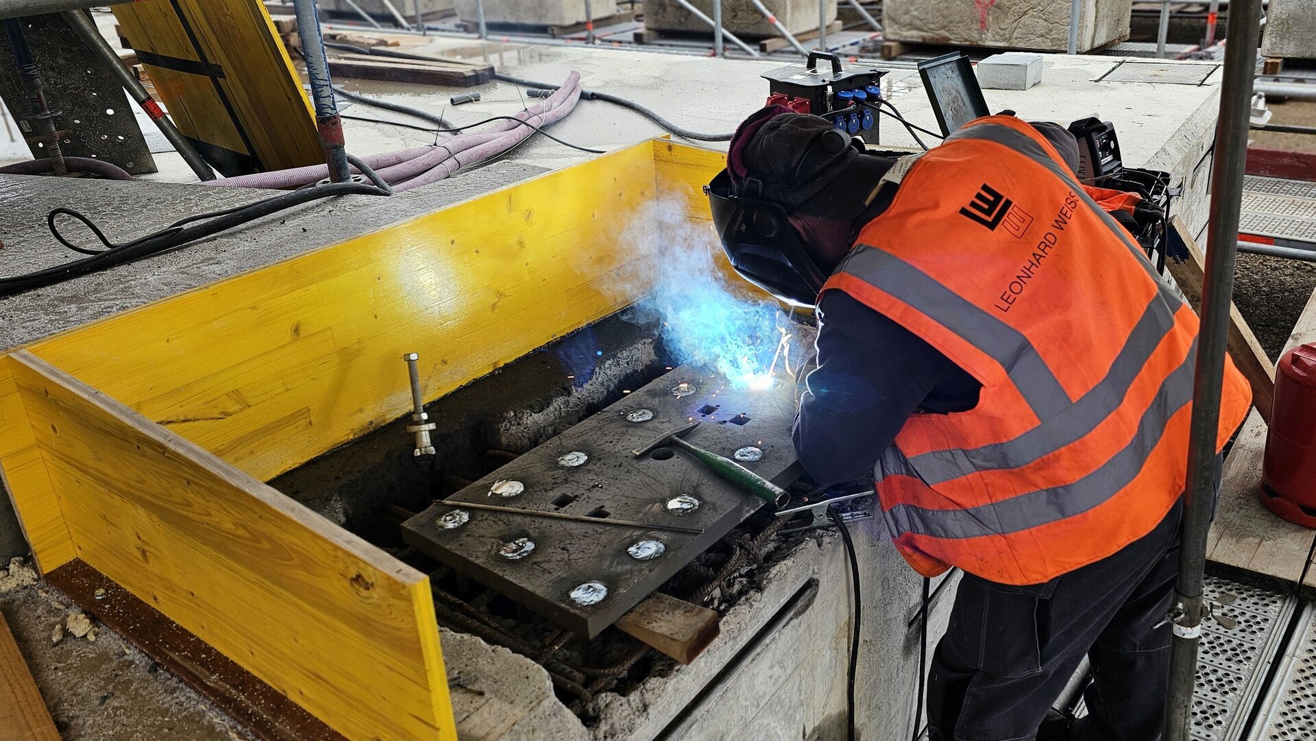 Auf einer Baustelle schweißt ein Arbeiter in orangefarbener Sicherheitsweste mit Schutzhelm und Handschuhen, umgeben von Holz und Werkzeugen.
