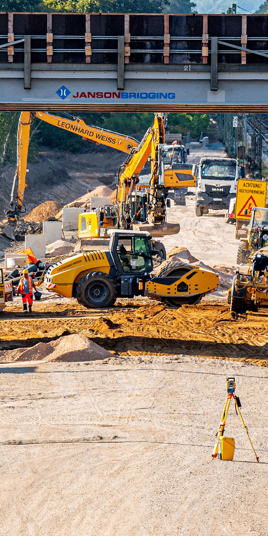 Auf der Baustelle sind verschiedene Baumaschinen aktiv, während im Hintergrund ein Brückenbauwerk und Gleise zu sehen sind.