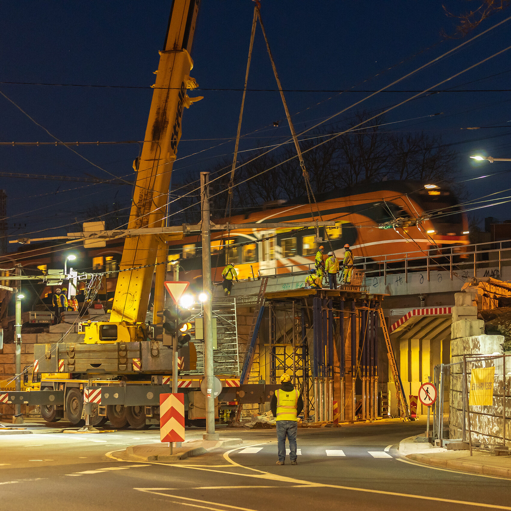 Bauarbeiten an einer Brücke bei Nacht, mit einem Zug, der schnell vorbeifährt, und Bauarbeitern, die konzentriert arbeiten.