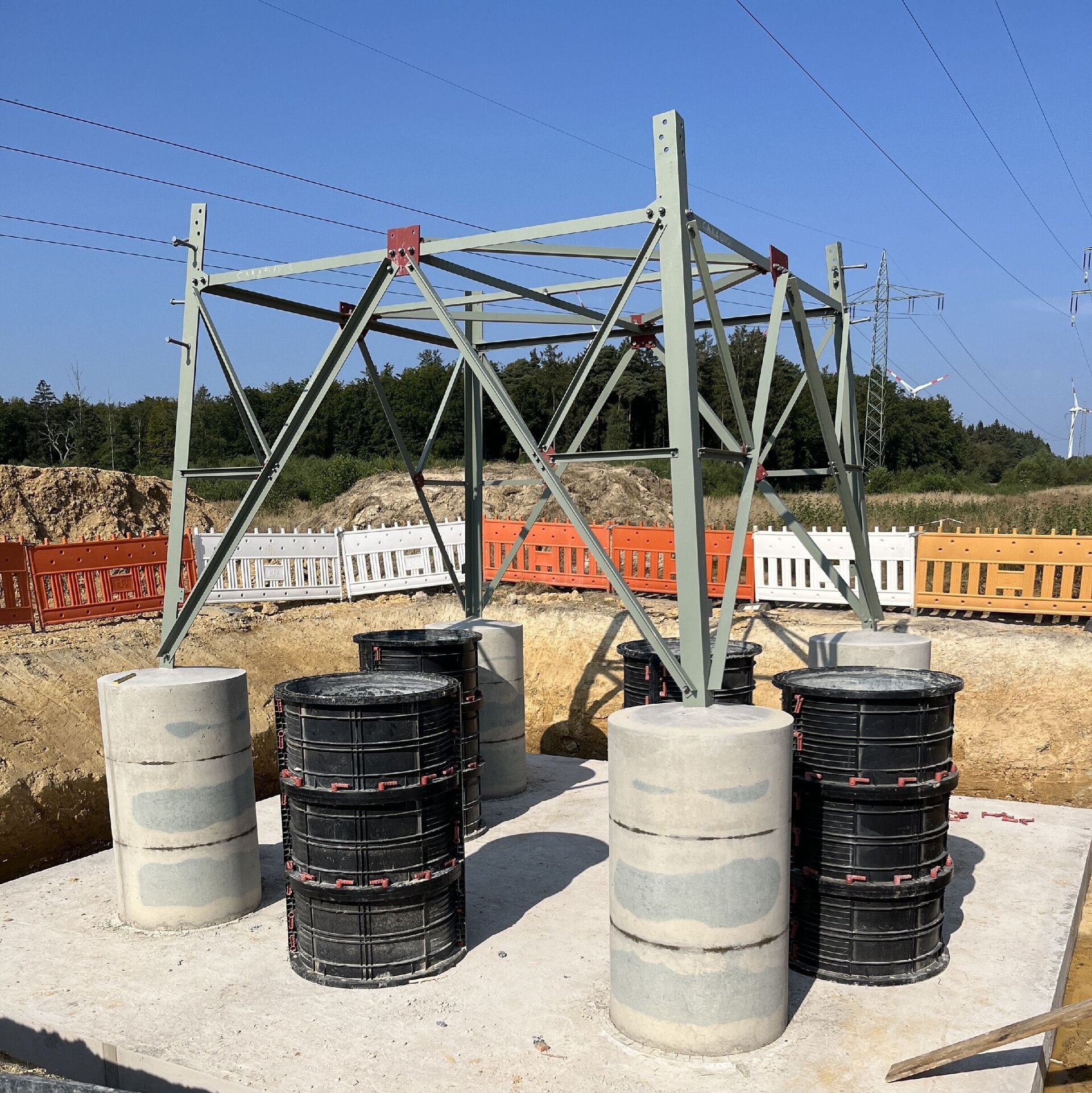 Das Bild zeigt auf einem abgesperrten Bereich die Basis eines Strommasten auf dem fertiggestellten Betonfundament, umgeben von Baustellenmaterialien.