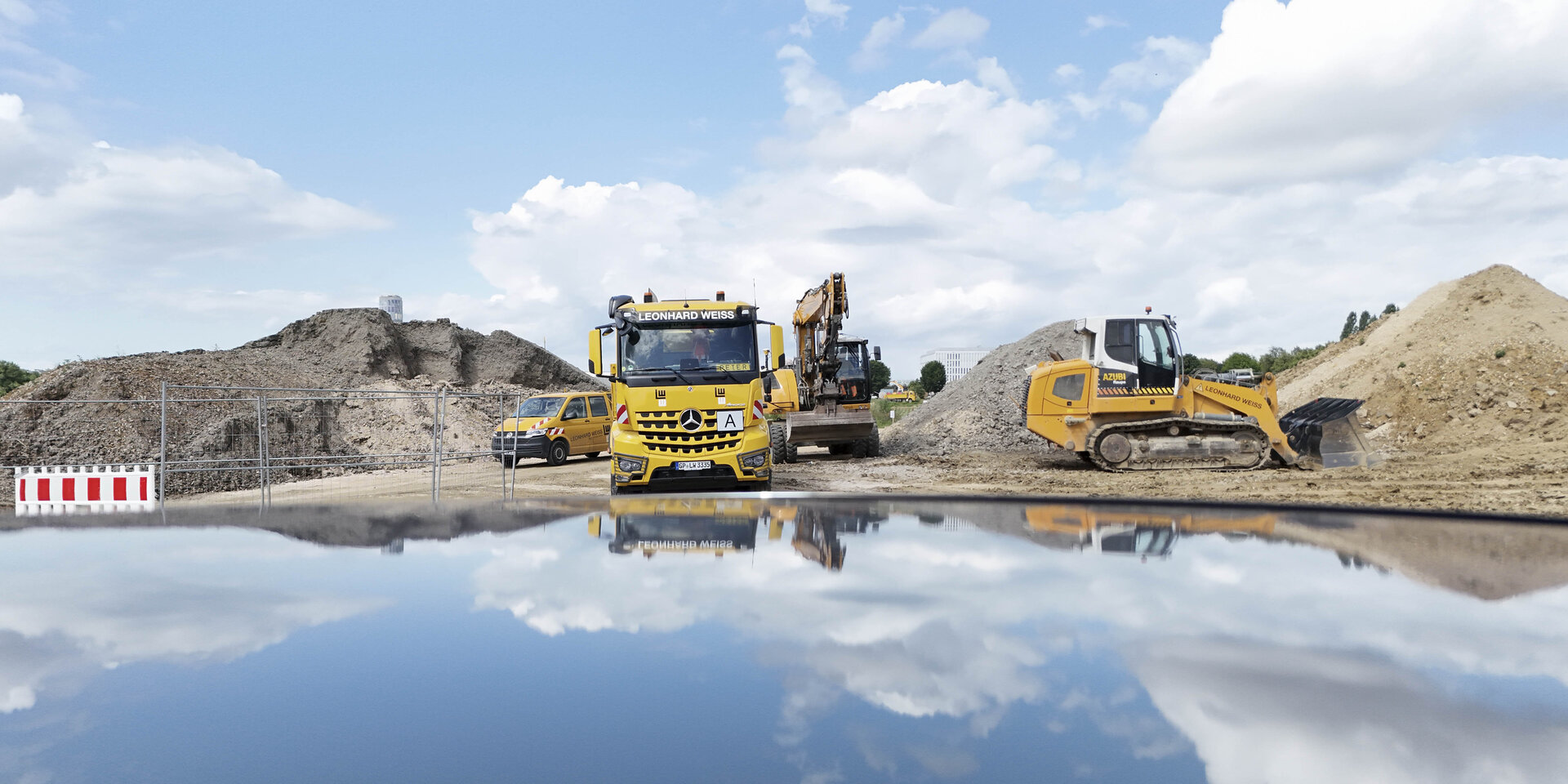 Gelbe Baugeräte stehen auf einer Baustelle, während sich der Himmel und Wolken im Wasser spiegeln.