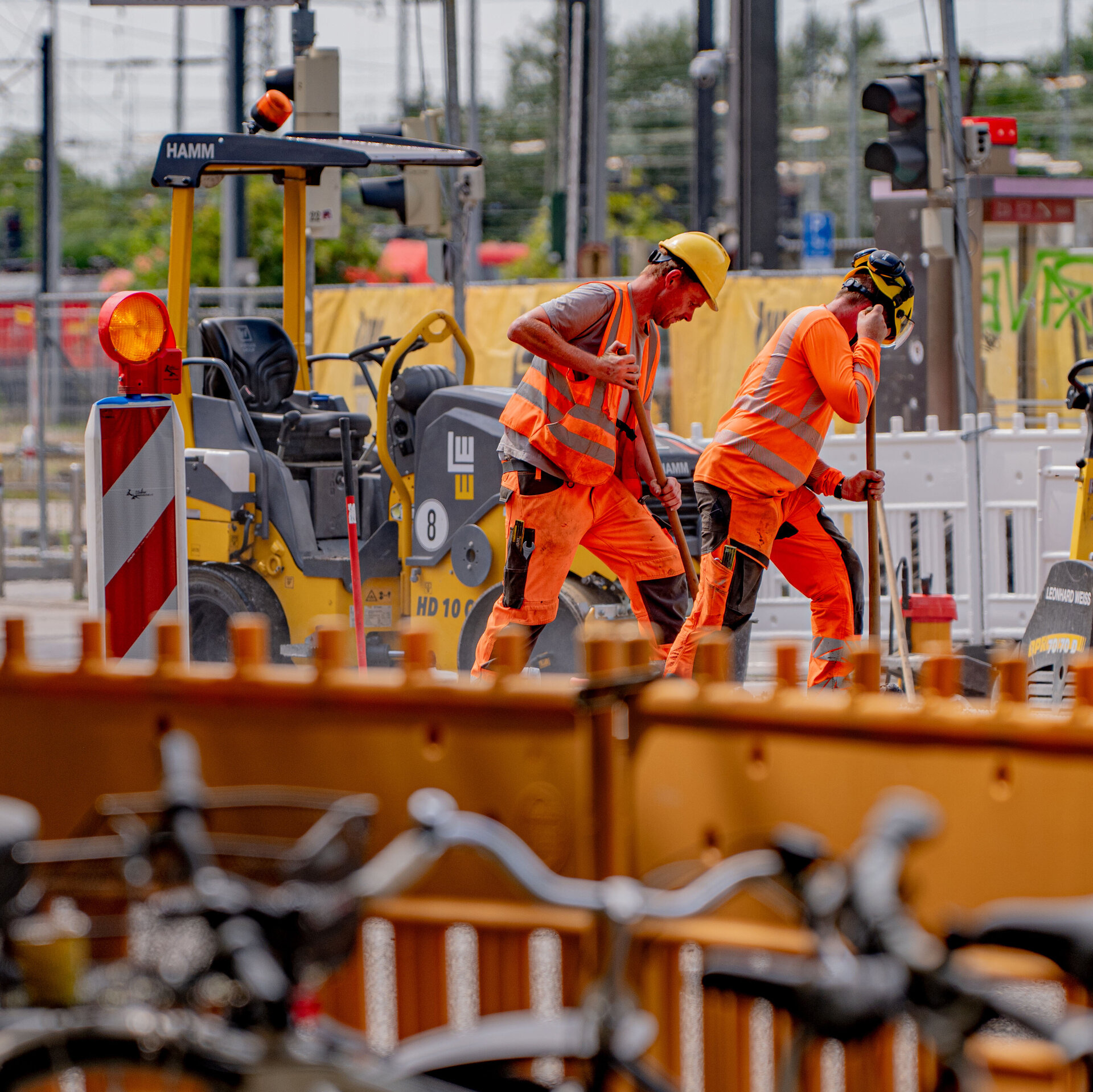 Zwei Bauarbeiter in leuchtend orangefarbener Kleidung arbeiten an einer Baustelle, während ein gelbes Baugerät im Hintergrund steht.