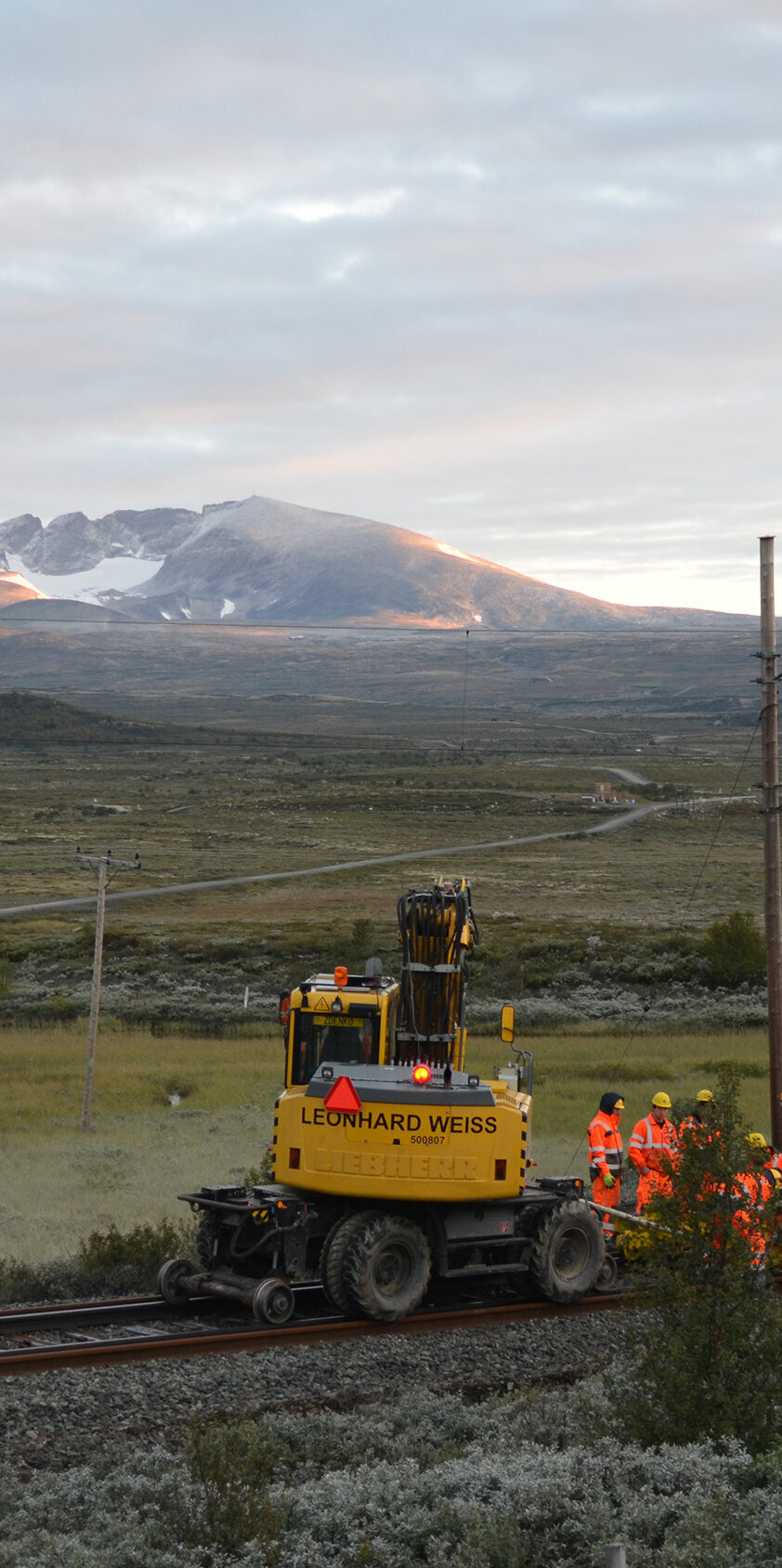 Ein Team von Bauarbeitern in leuchtenden Anzügen führt Arbeiten an den Bahngleisen durch, während die Sonne hinter den Bergen aufgeht.