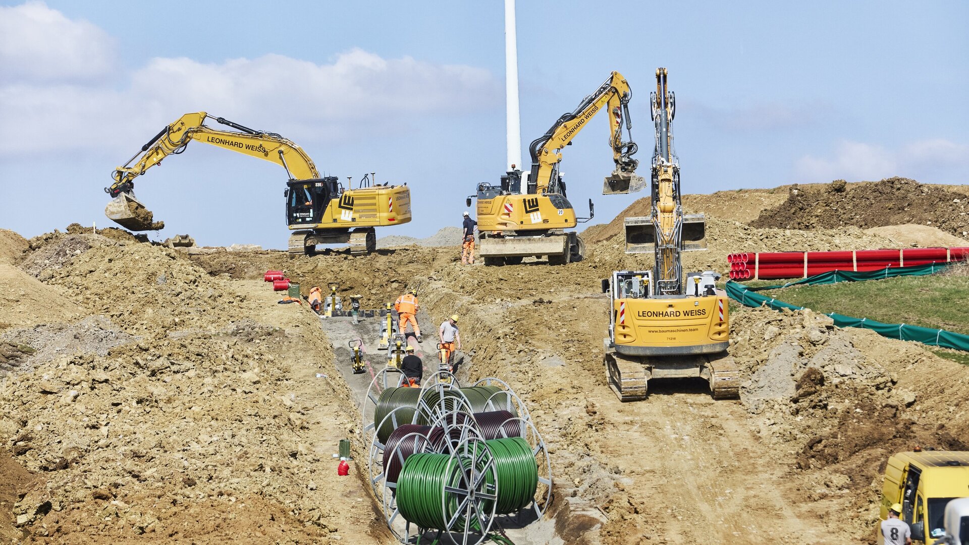 Gelbe Bagger heben Erde auf einer Baustelle, während Arbeiter in orangefarbener Kleidung Kabel und Rohre vorbereiten.