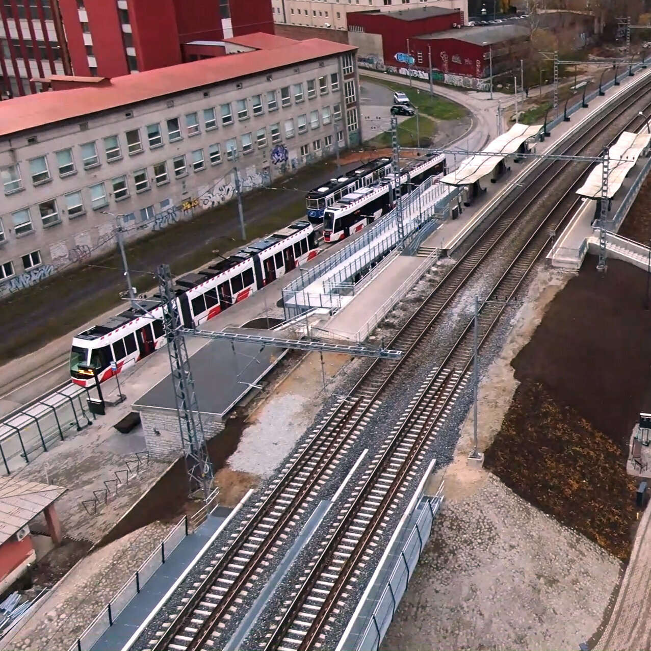 Die Szene zeigt eine Straßenbahnlinie mit mehreren Zügen, die an einer neuen Haltestelle in einer urbanen Umgebung hält.