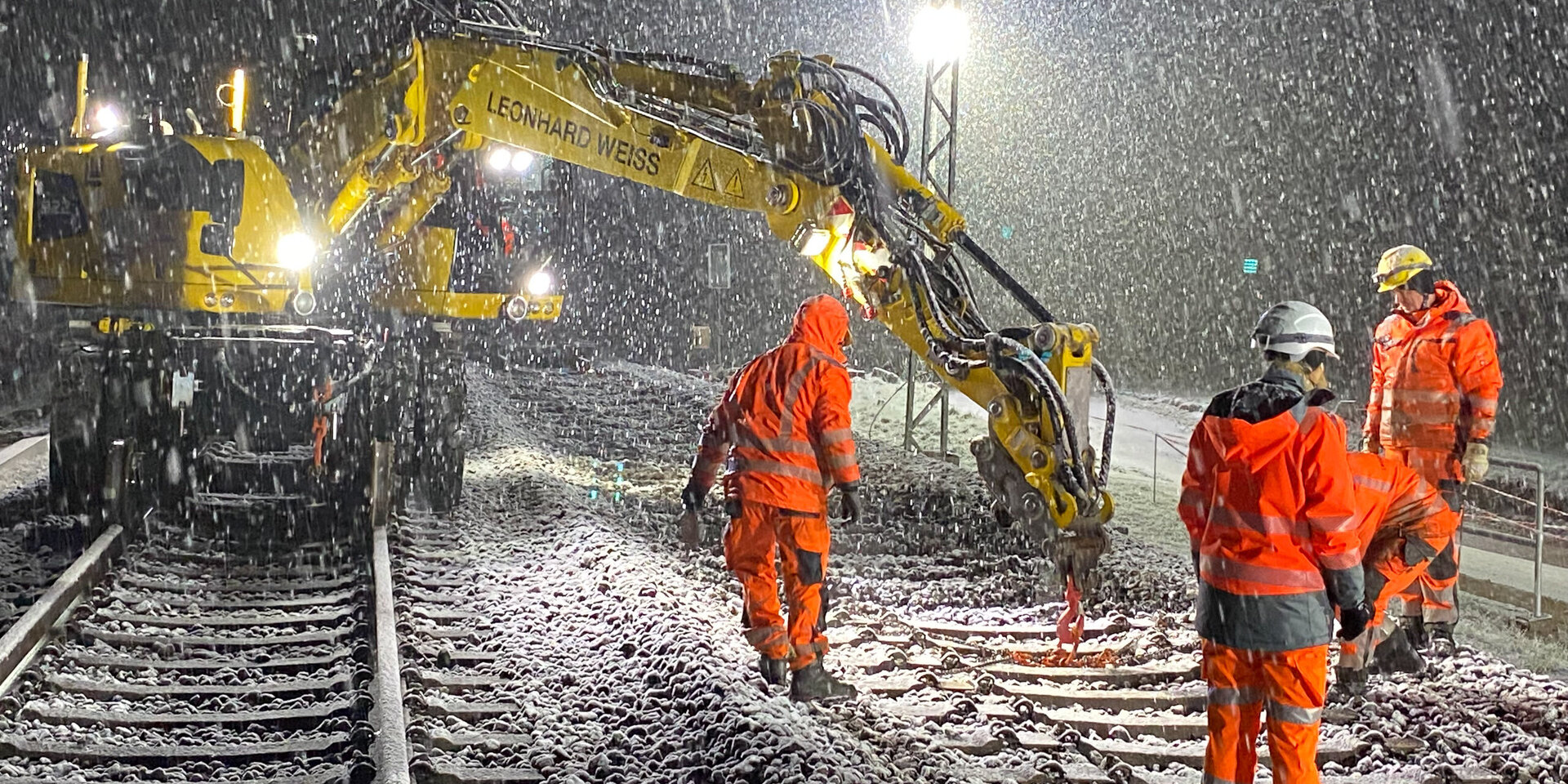 In der Nacht, während es schneit, sind Arbeiter mit Helmen und Warnkleidung mit einer Maschine an den Bahngleisen beschäftigt.