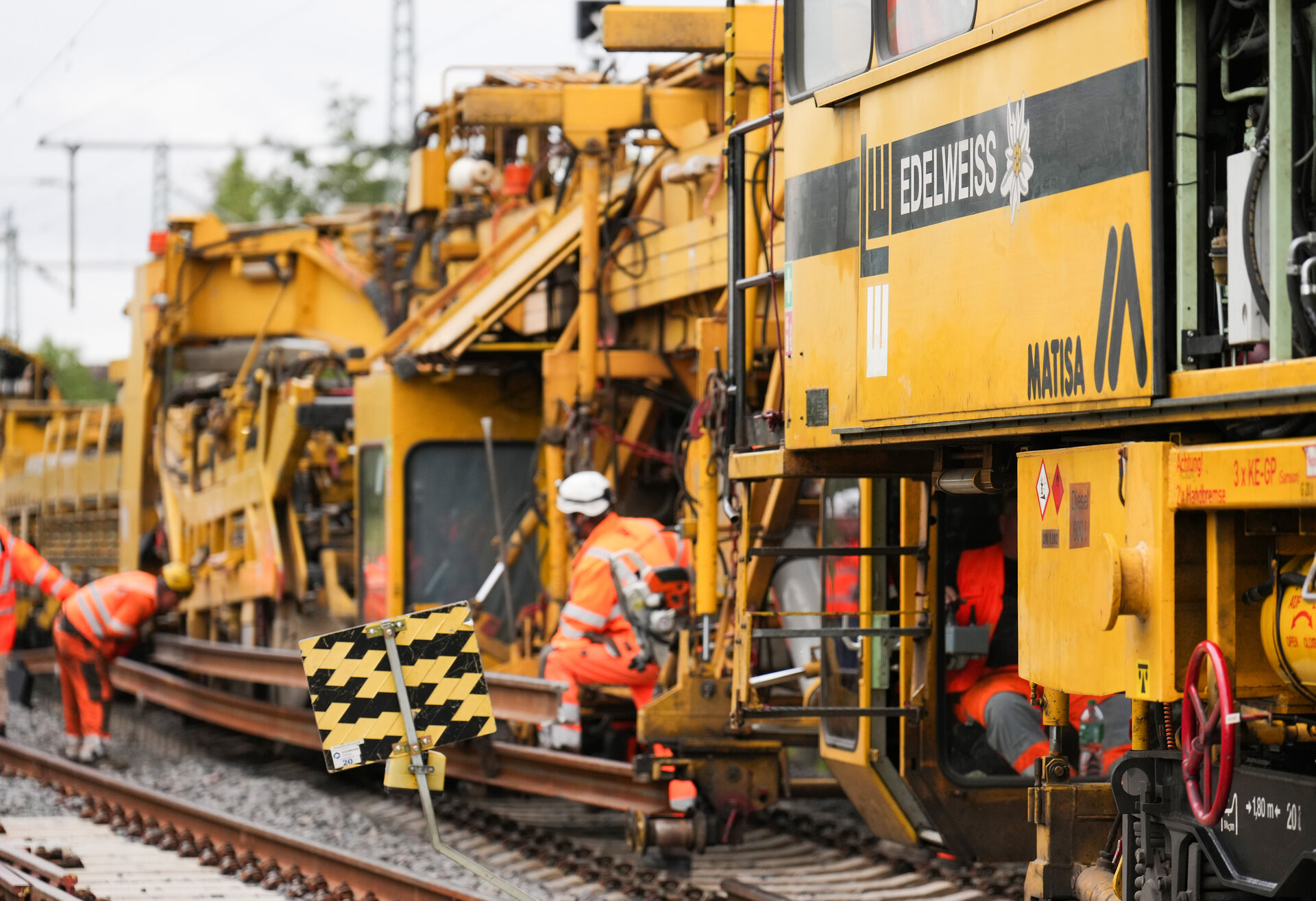 Arbeiter in leuchtend orangefarbener Kleidung führen Wartungsarbeiten an einer Bahngleisanlage mit schweren Maschinen durch.