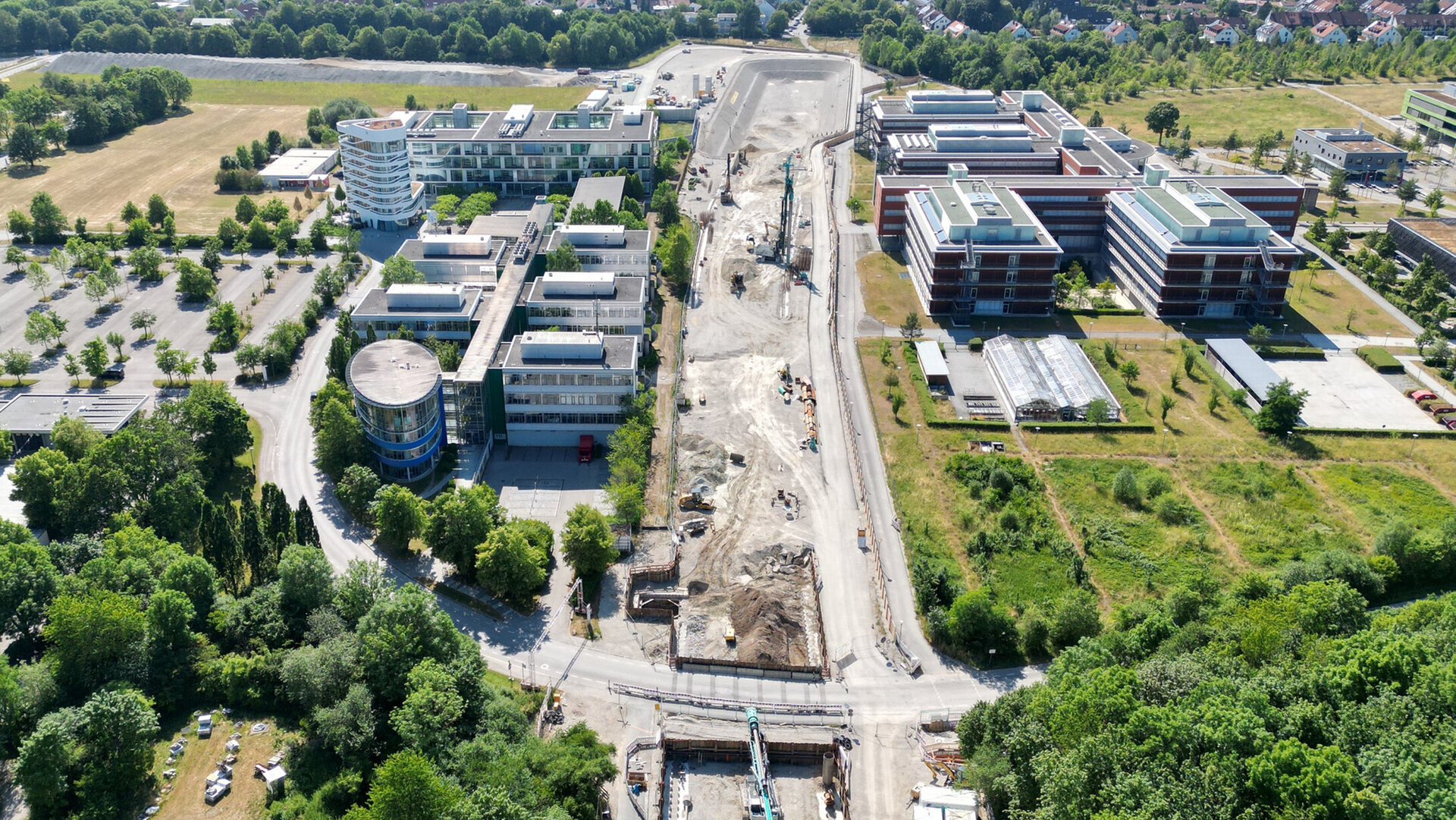 Im Bild sind moderne Bürogebäude und eine Baustelle zu sehen, umgeben von viel Grün und einer Waldlandschaft im Hintergrund.