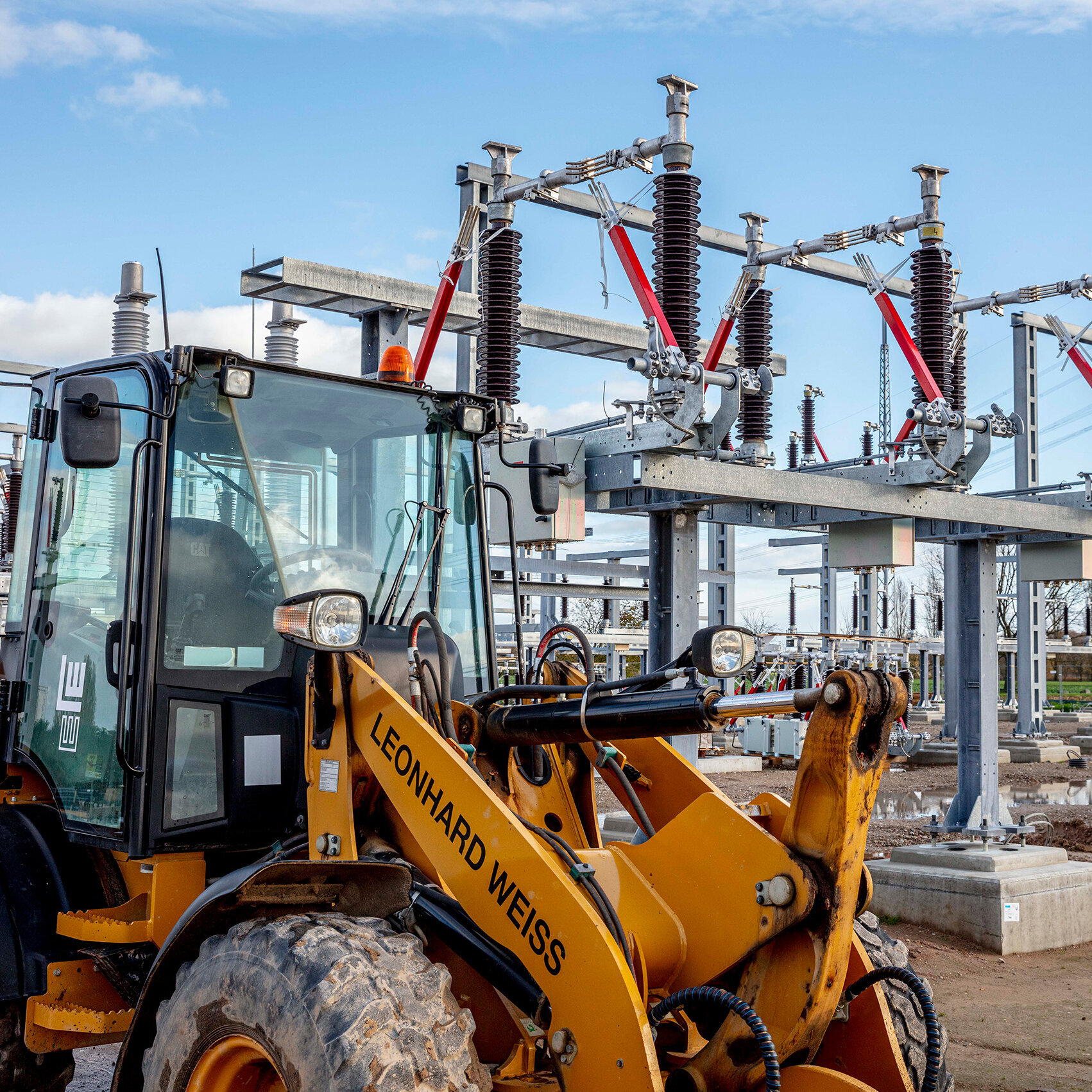 Ein gelber Bagger steht vor einer modernen Umspannstation mit hohen Stromleitungen und großen Transformatoren im Hintergrund.