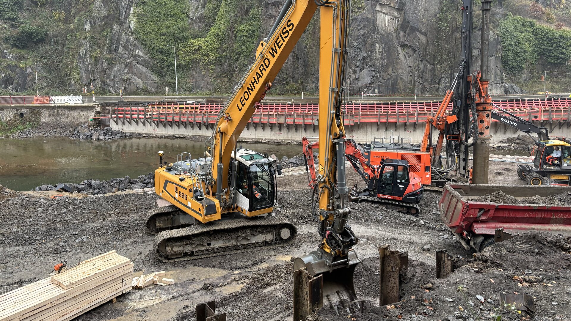 Im Vordergrund mehrere Bagger beim Aushub von Erde für die Installation von Holzverstrebungen auf der Baustelle im Bereich um die „Bunte Kuh“, während im Hintergrund eine rote Brücke und steile Felsen sichtbar sind.