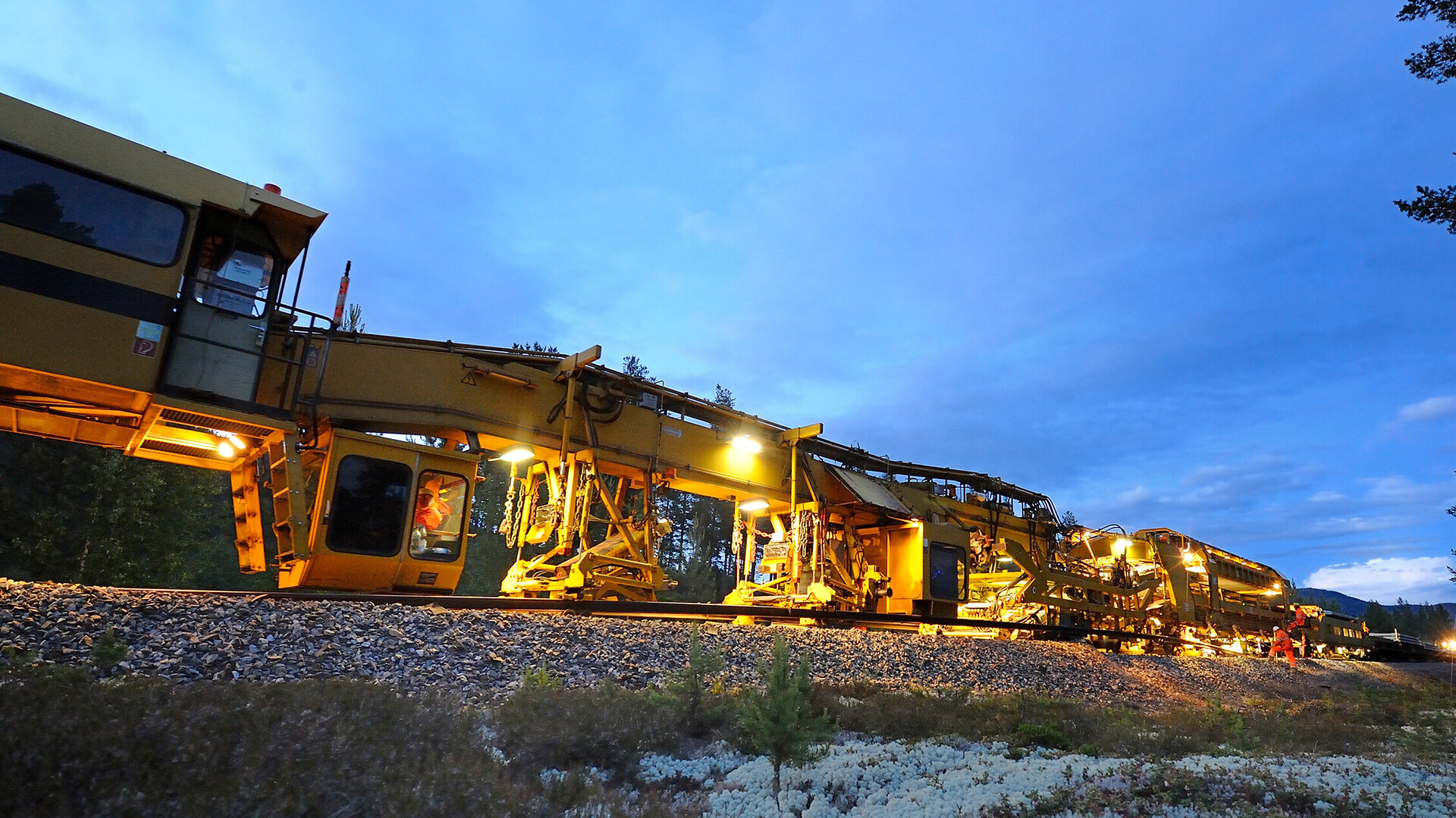In der Dämmerung ist eine gelbe Gleisbaumaschine zu sehen, die mit Scheinwerfern die Schienen bearbeitet, umgeben von Natur.