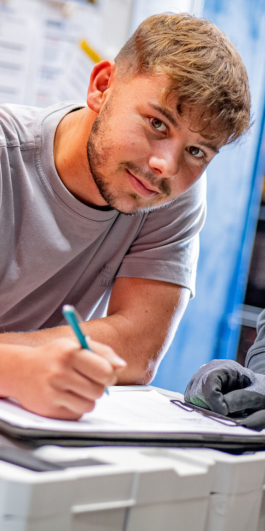 Ein junger Mann in einem grauen T-Shirt schreibt konzentriert auf einem Dokument, während andere Personen ihm über die Schulter schauen.