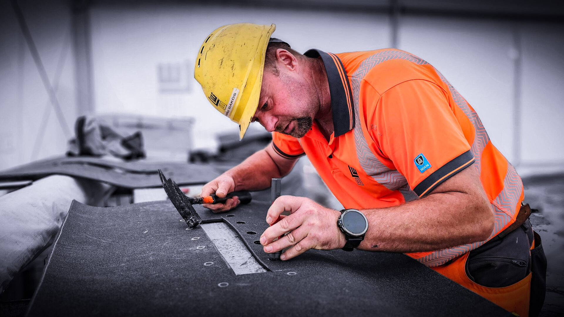 Ein Handwerker in orangefarbener Arbeitskleidung schneidet und vorbereitet eine Schweißbahn mit einem Hammer und einem Werkzeug.