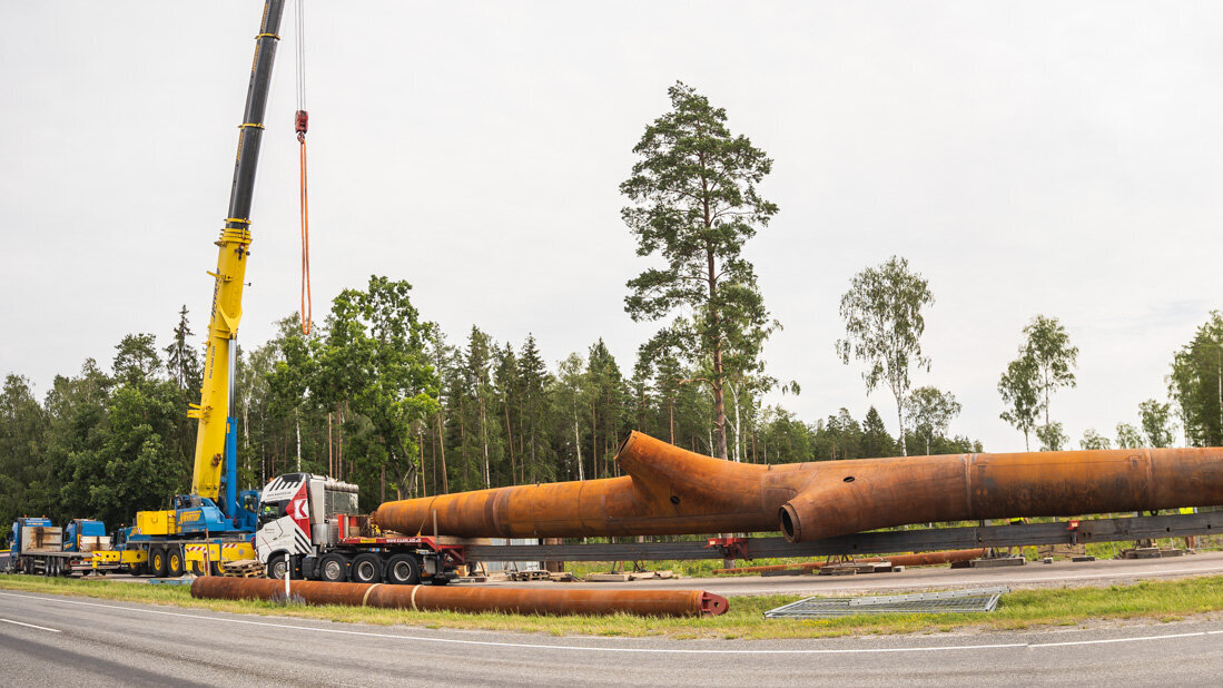 Auf einer Baustelle wird ein massives, rostiges Rohr von einem Kran angehoben, während ein Lkw in der Nähe parkt und Bäume im Hintergrund stehen.