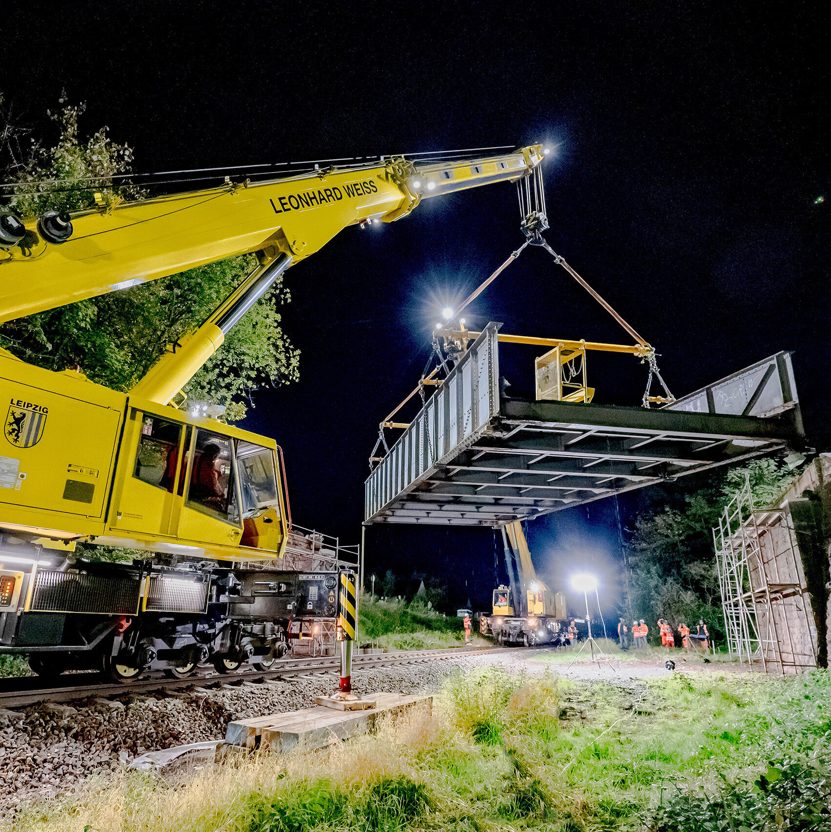 Bei Nacht wird eine Brückenkonstruktion von einem Kran über die Gleise gehoben, umgeben von beleuchteten Baustellen und Bäumen.