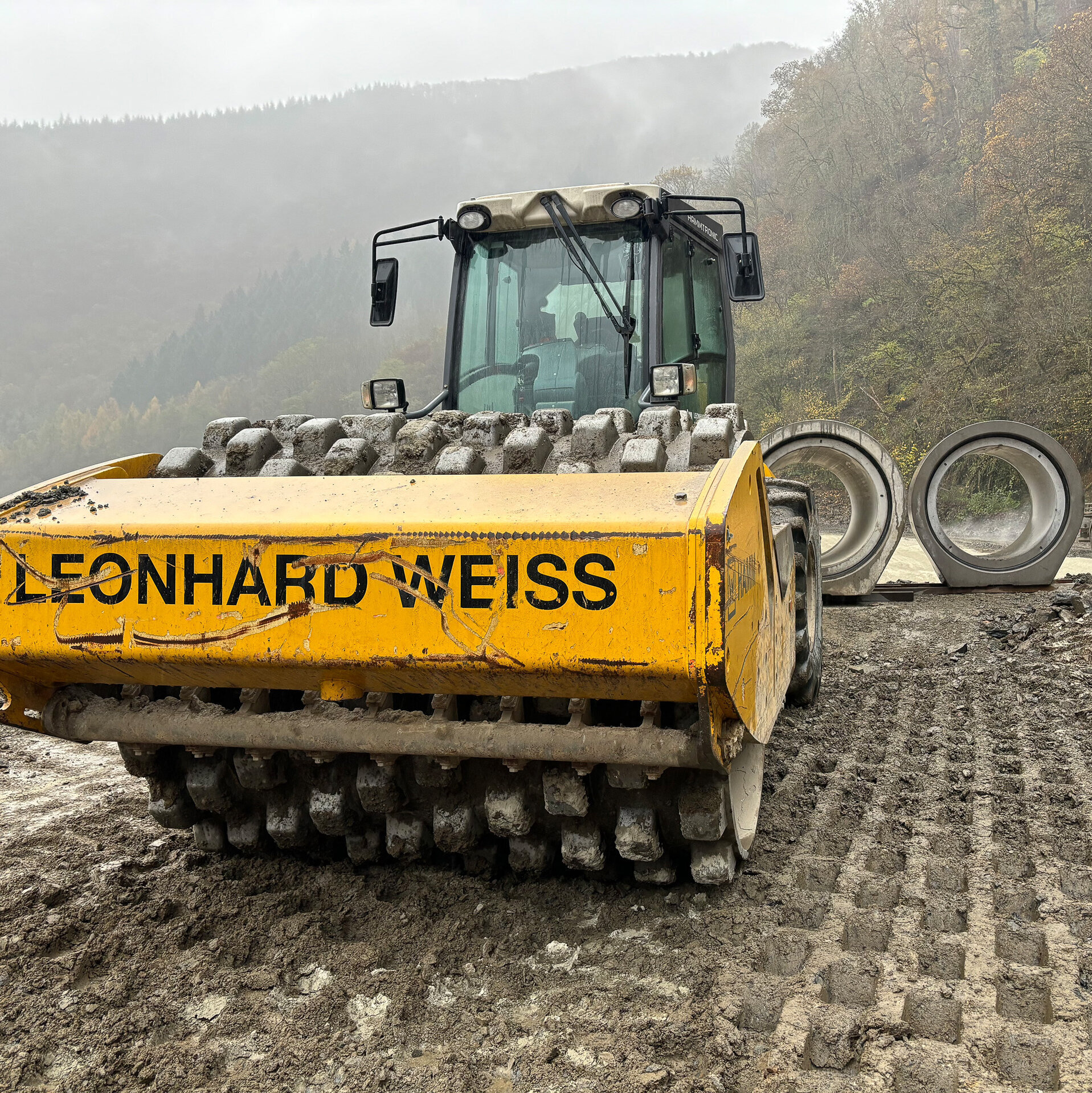 Ein gelber Walzenfahrer steht im Vordergrund, während große Betonrohre im Hintergrund auf einer Baustelle liegen.