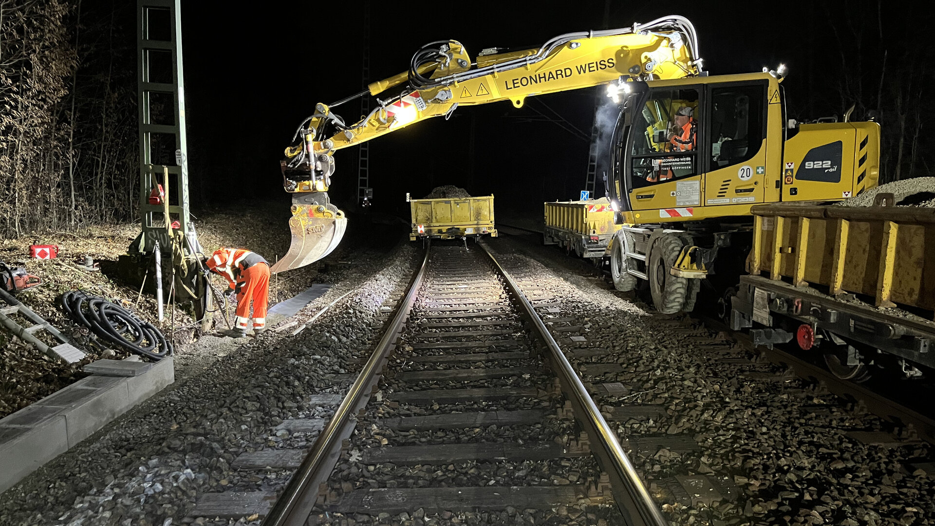 Nachtaufnahme von Gleisbauarbeiten beim Einheben von Betonkabelkanalteilen mit Zwei-Wege-Bagger.