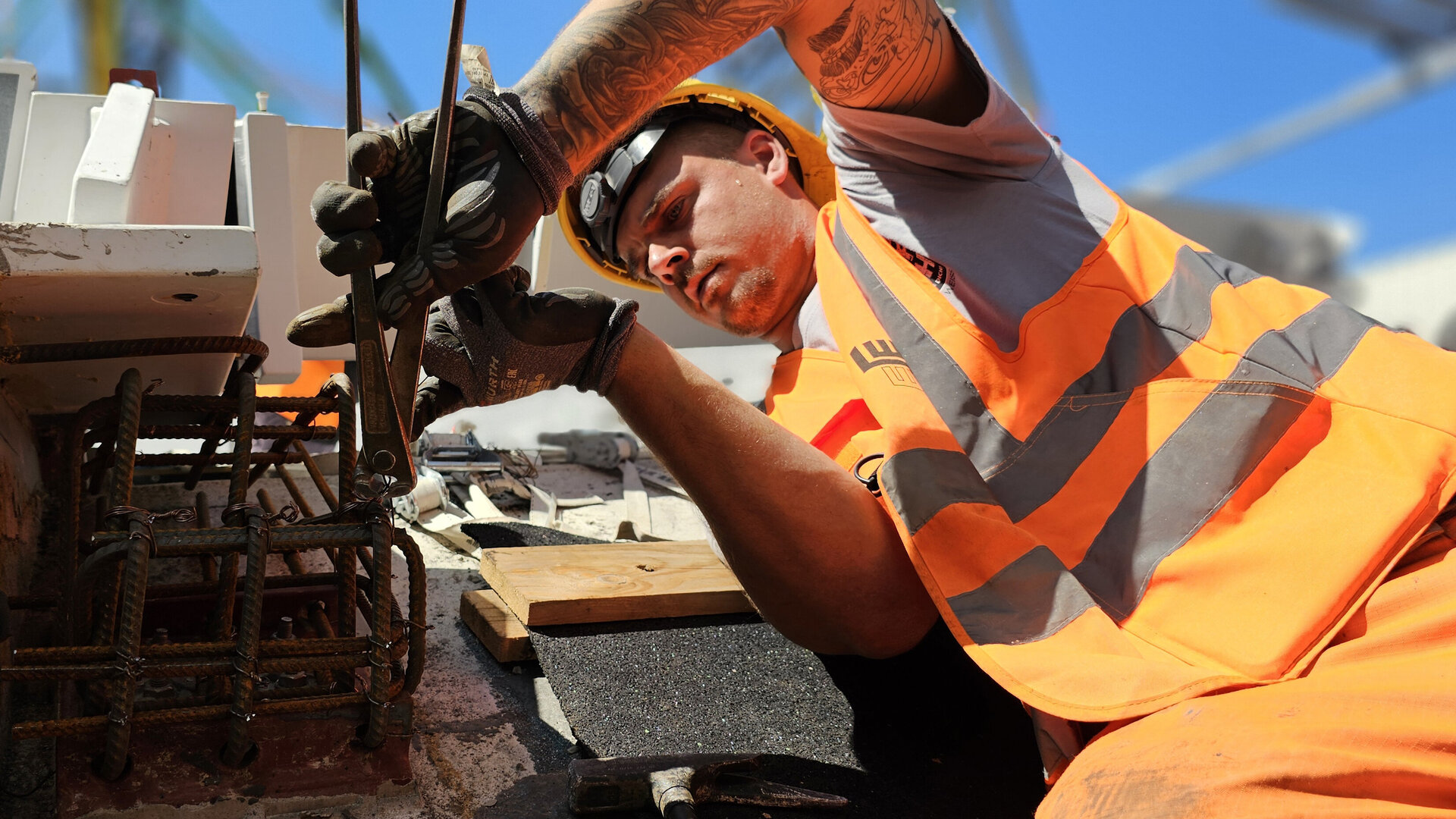 Ein Bauarbeiter in orangefarbener Sicherheitskleidung bindet im Freien mit einer Zange einen Bewehrungskorb.