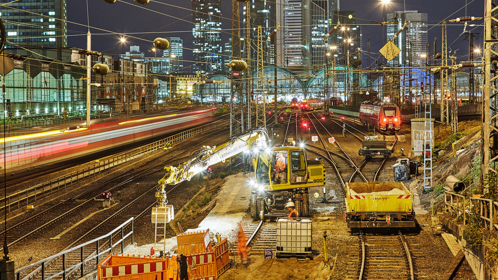 The skyscrapers of Frankfurt glow in the background as a train with red lights passes by on the tracks.