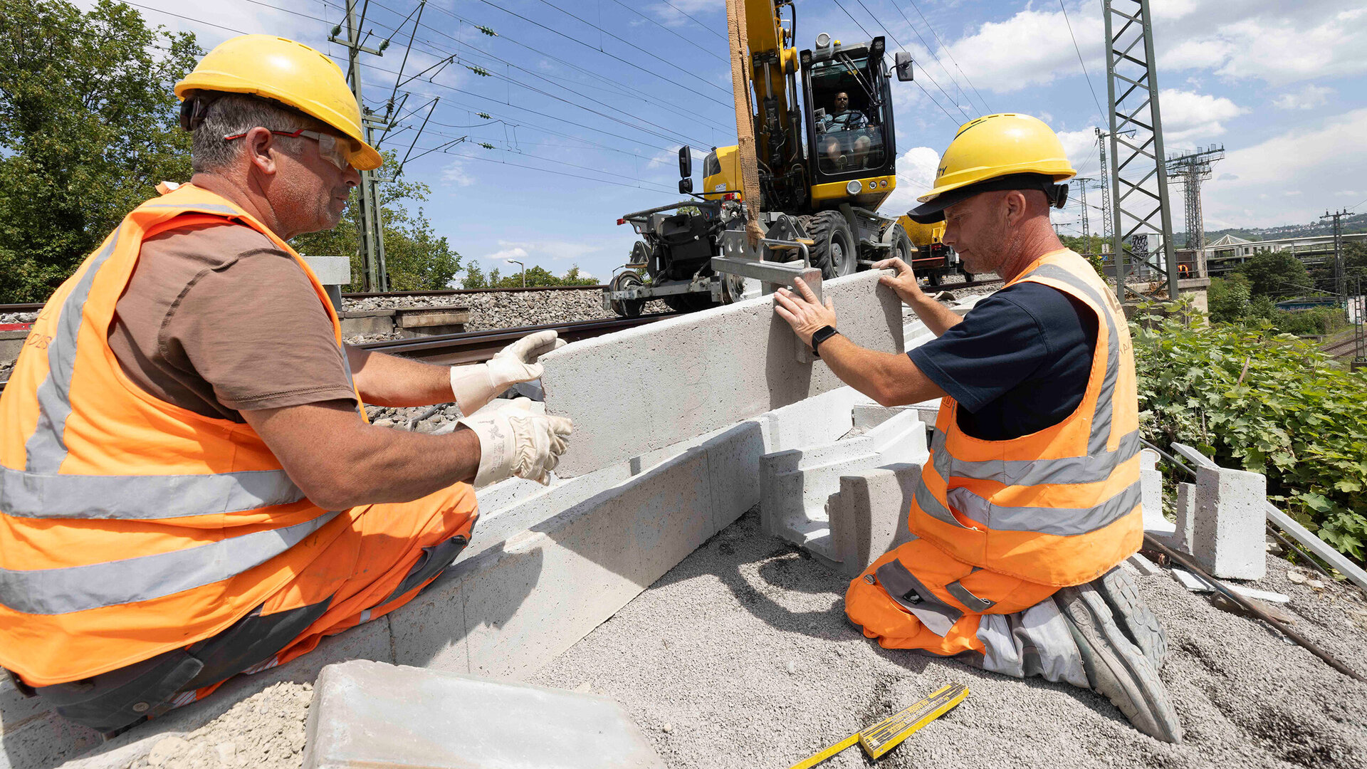 Zwei Bauarbeiter in orangefarbener Sicherheitskleidung arbeiten an einer Baustelle neben den Bahngleisen und heben Betonblöcke ins Gleisbett.