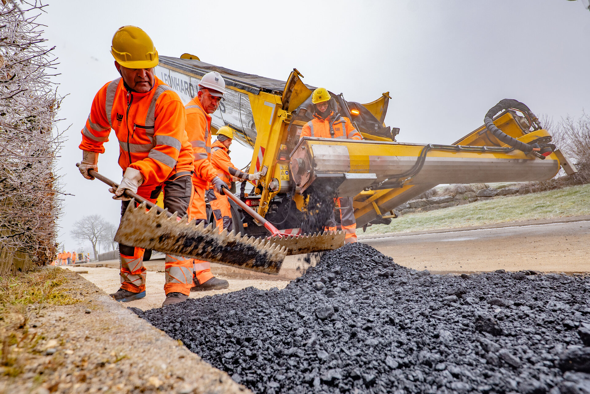 Eine Gruppe von Bauarbeitern nutzt Schaufeln, um frischen Asphalt zu verteilen, während eine große Maschine Asphalt abgibt.