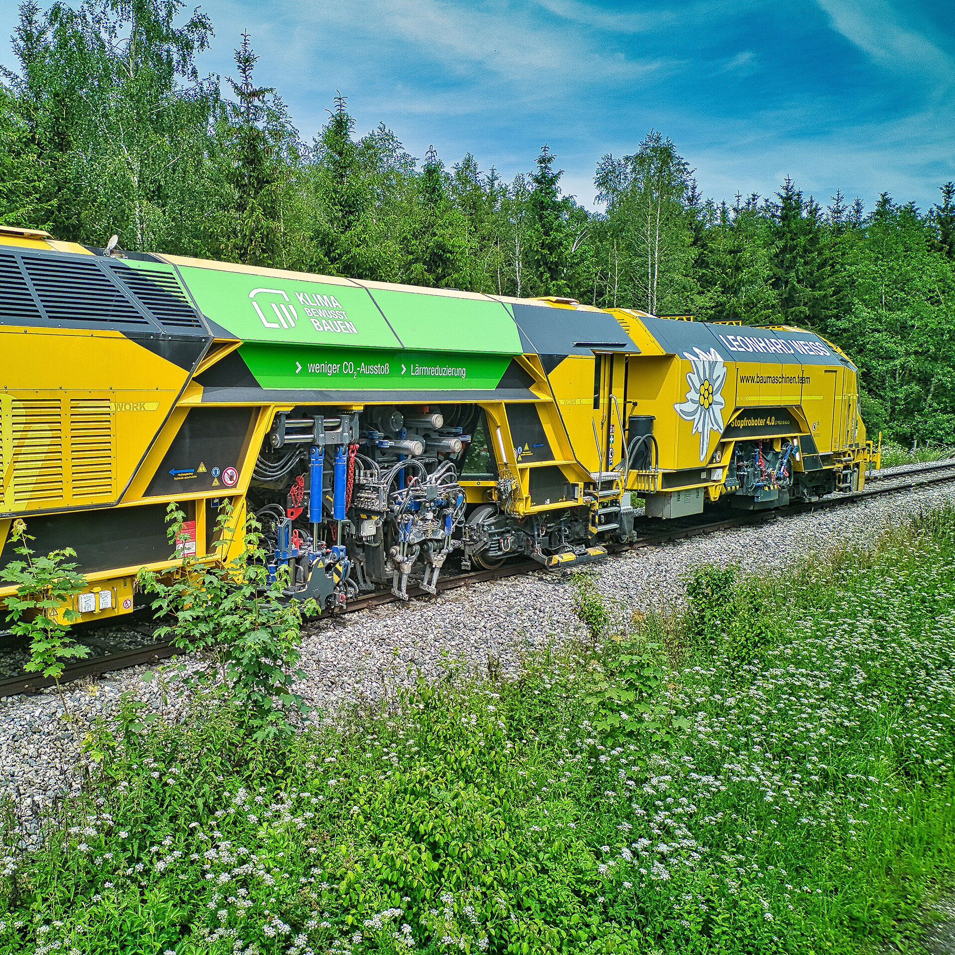 Ein gelber Gleisbauzug steht auf einer Schotterstrecke, umgeben von üppigem Grün und blühenden Pflanzen.