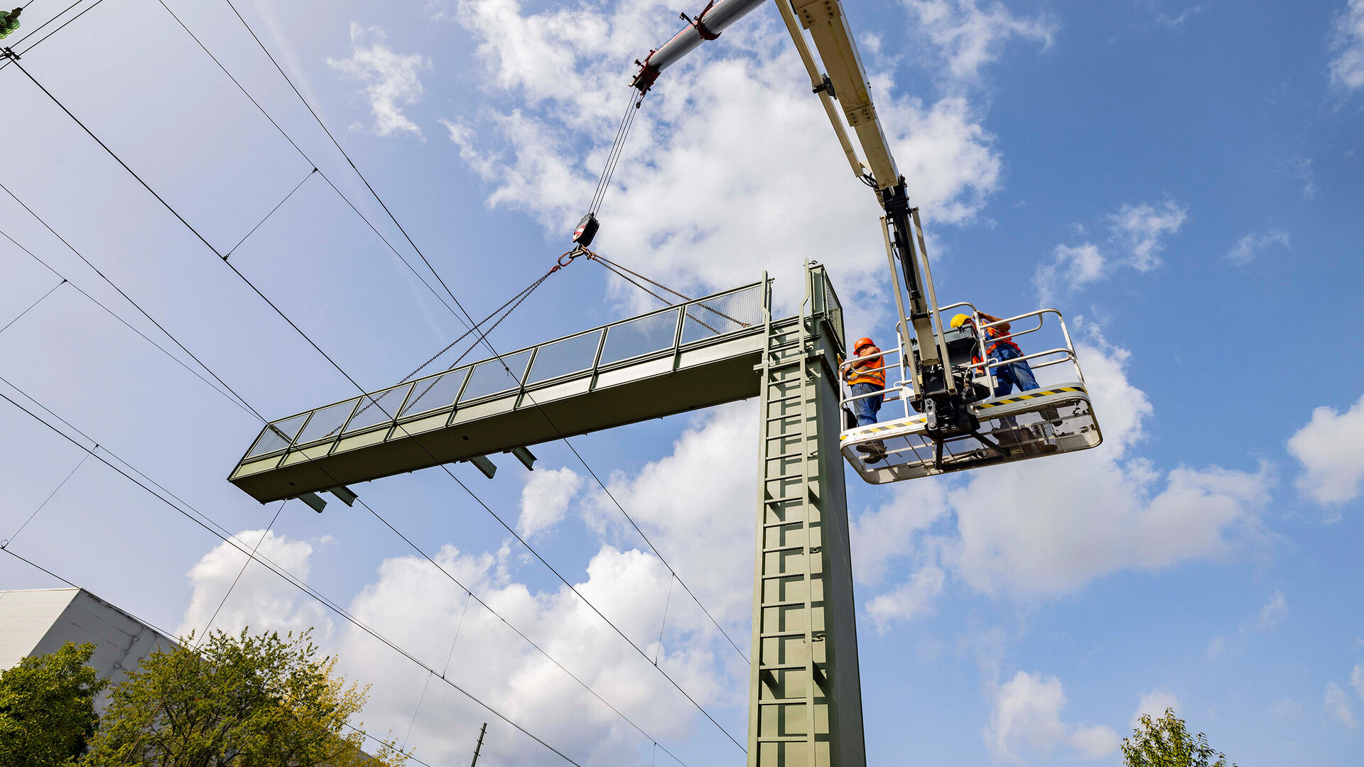 Zwei Bauarbeiter in orangefarbenen Helmen stehen auf einer Hebebühne, während ein Signalausleger mit einem Kran installiert wird.