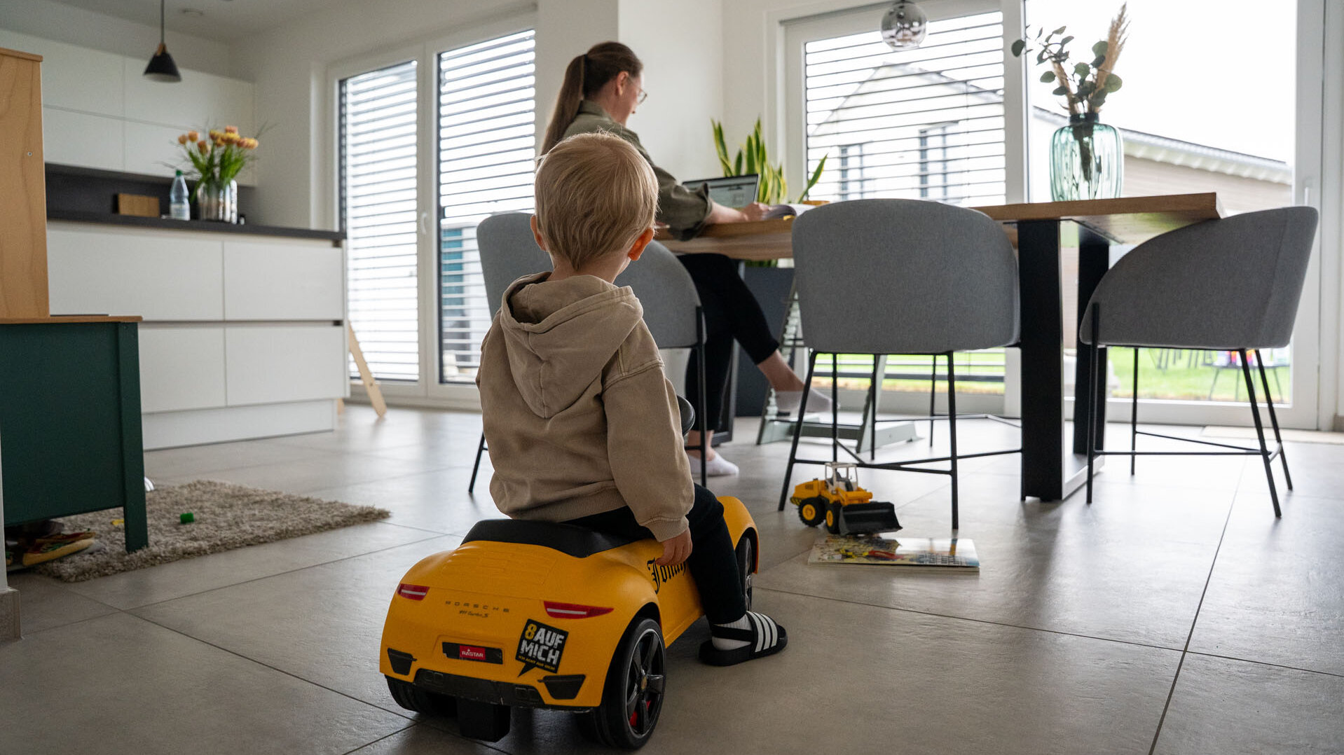 Ein Junge auf einem gelben Spielzeugauto beobachtet seine Mutter, die am Esstisch mit einem Laptop arbeitet.