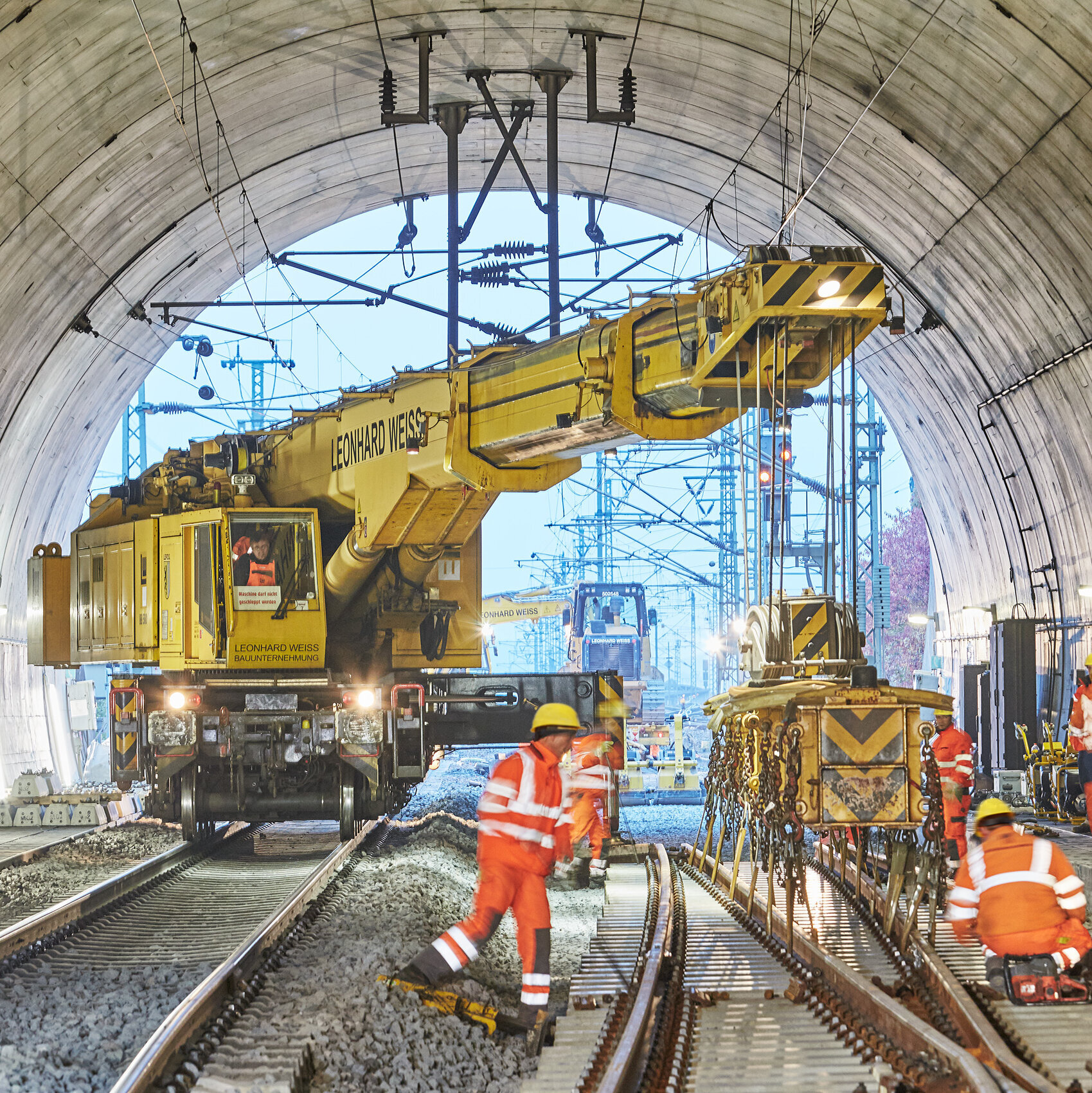 Ein gelber Kran hebt Ausrüstung in einem Tunnel, während Arbeiter an den Gleisen beschäftigt sind und Sicherheitshelme tragen.