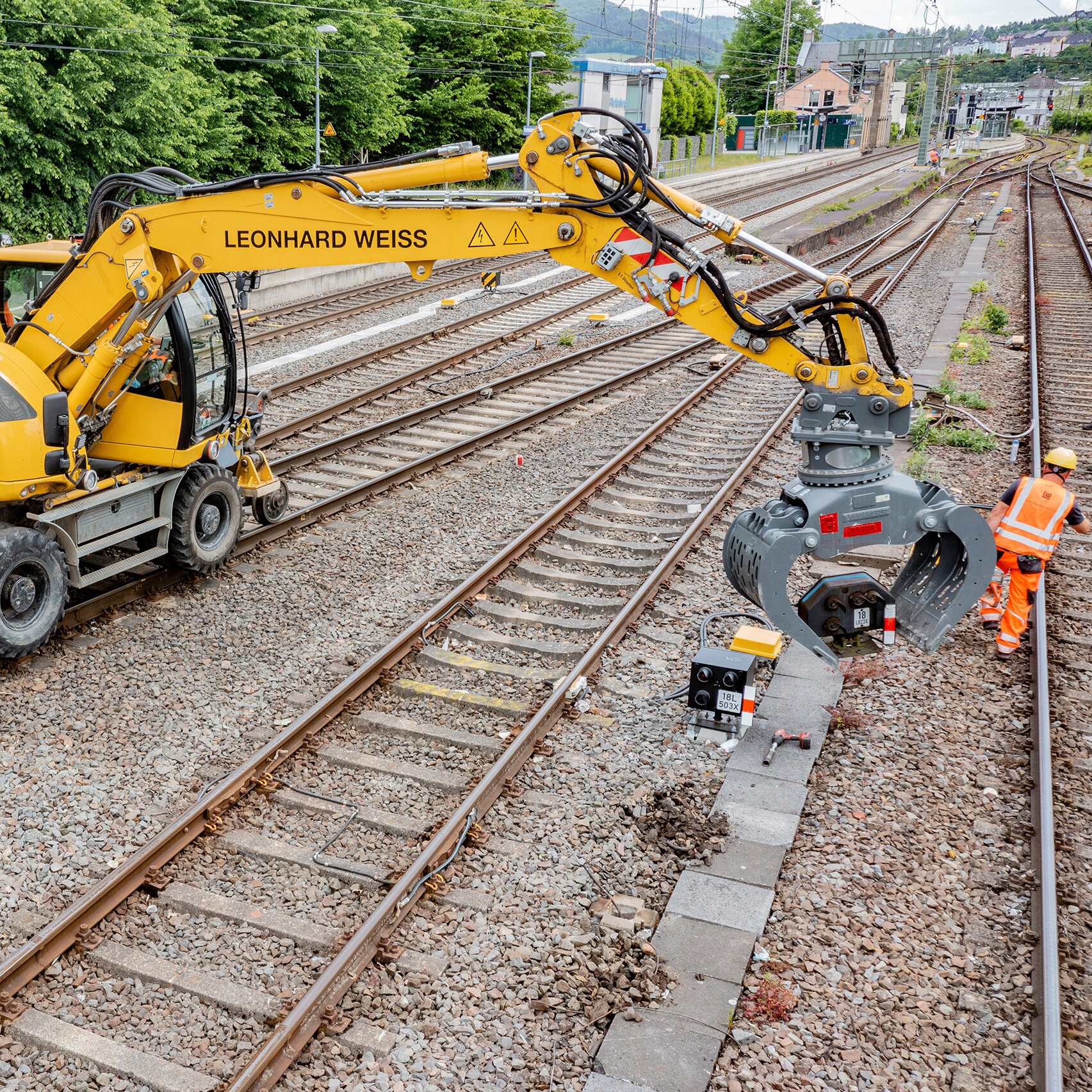Ein Gleisbauarbeiter in orangefarbener Sicherheitskleidung steht neben einem gelben Bagger, der an den Schienen arbeitet.