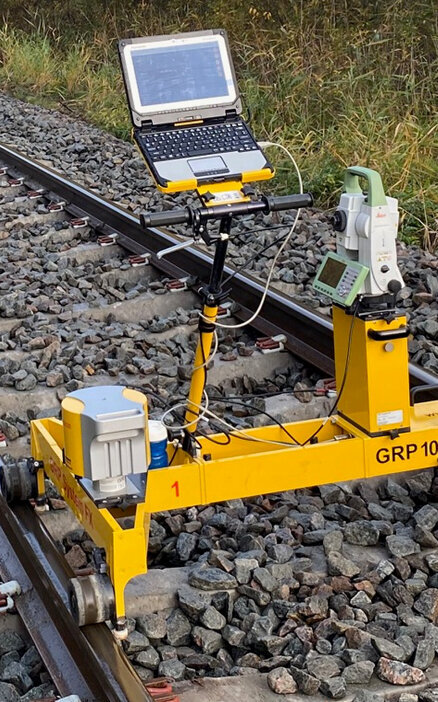 Ein gelbes Gleismessgerät steht auf Schotter zwischen den Bahngleisen, ausgestattet mit einem Laptop und verschiedenen Sensoren.