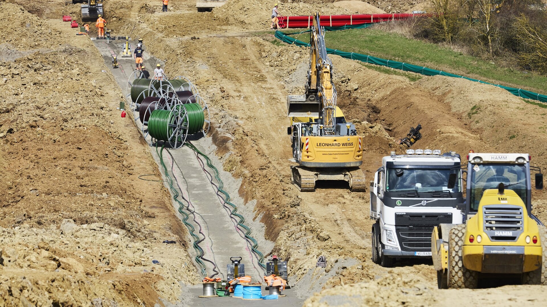 Bauarbeiter verlegen Kabel auf einer Baustelle, während Bagger und Lkw im Hintergrund für den Transport von Materialien sorgen.