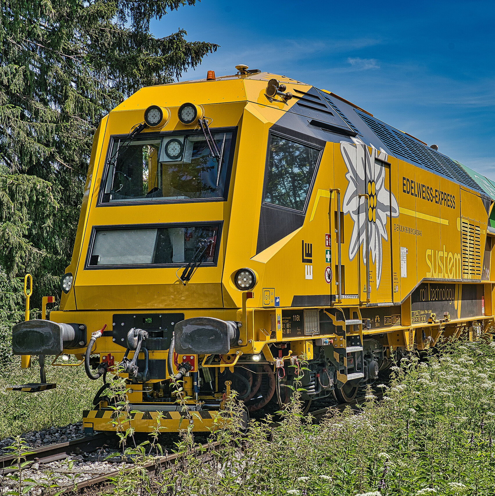 Ein gelber Gleisbauzug mit dem Namen 'Edelweiss-Express' steht auf einem Schottergleis, umgeben von grünen Bäumen und Pflanzen.