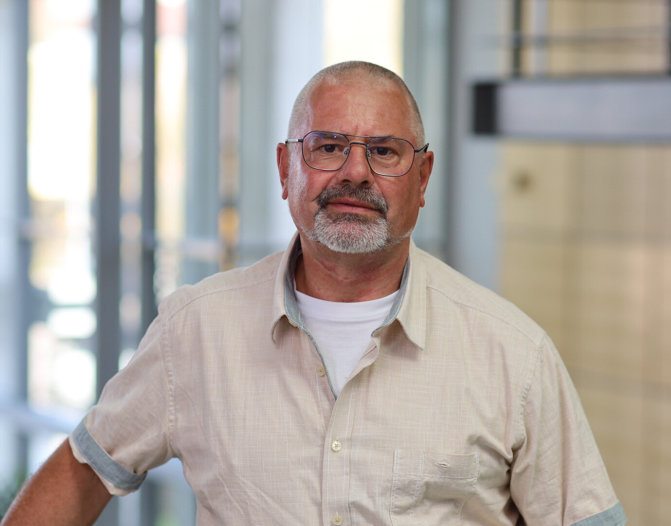 Der Mann mit einem kurzen grauen Bart und Brille lächelt leicht, während er in einem modernen, lichtdurchfluteten Raum steht.