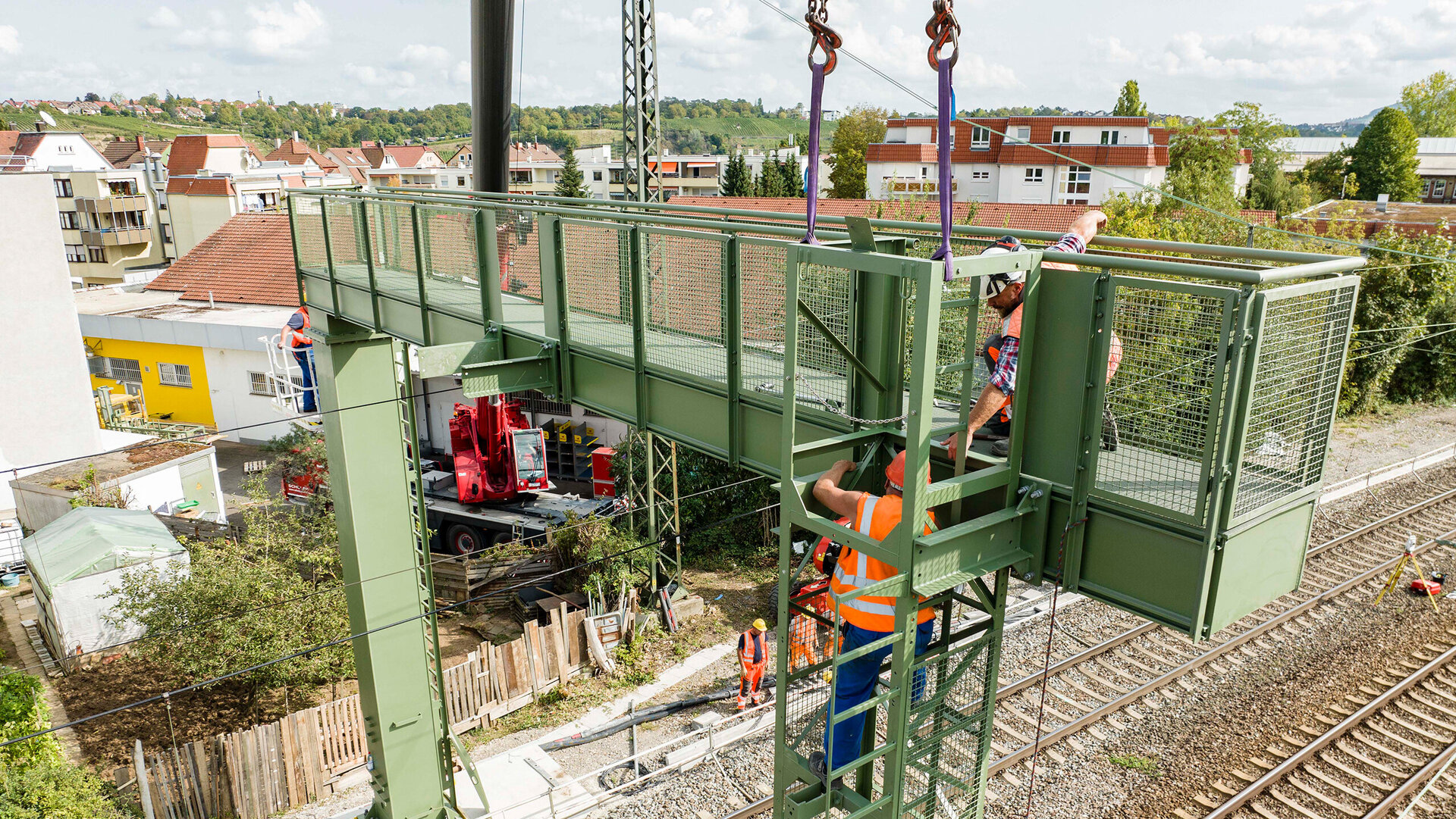 Arbeiter in Sicherheitswesten montieren eine grüne Plattform über den Bahngleisen, während ein Kran im Hintergrund sichtbar ist.