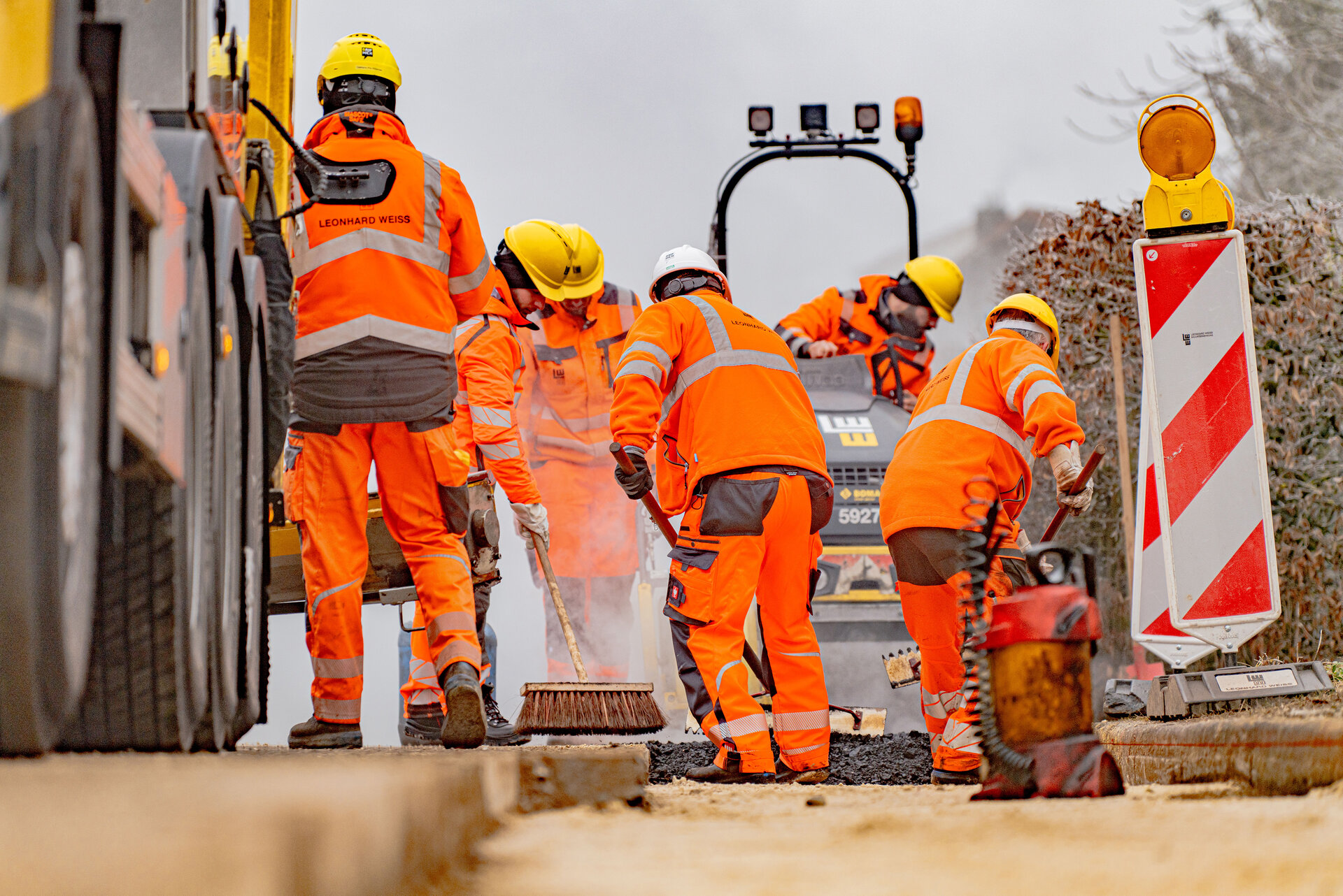 Fünf Bauarbeiter in leuchtend orangefarbener Kleidung arbeiten an einer Straßenbau-Stelle, während ein sechster Arbeiter die Walzmaschine im Hintergrund bedient.